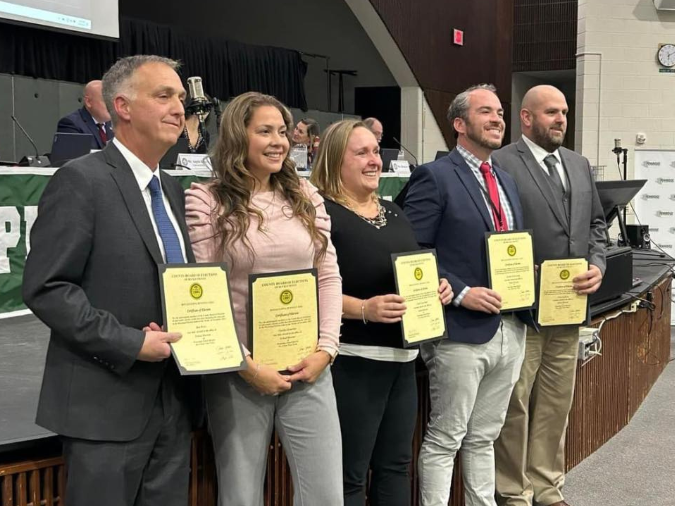 Bradley Merkl-Gump holding an award certificate during a school board meeting in a large room with a stage, a banner, and other people seated in the background.