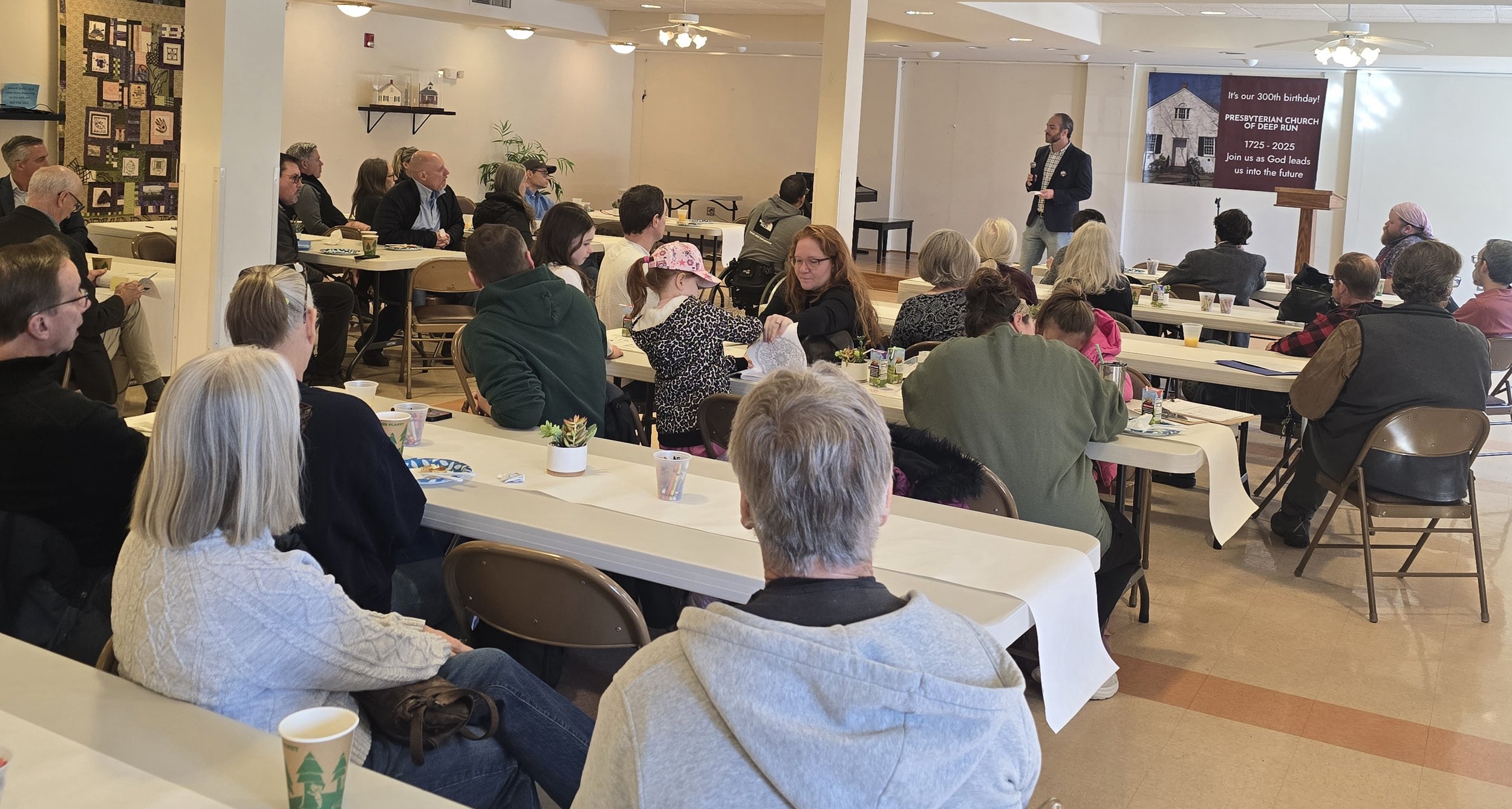A diverse group of people attending a community event or seminar in a hall, with Pennsylvania senate candidate Bradley Merkl-Gump standing at a podium.
