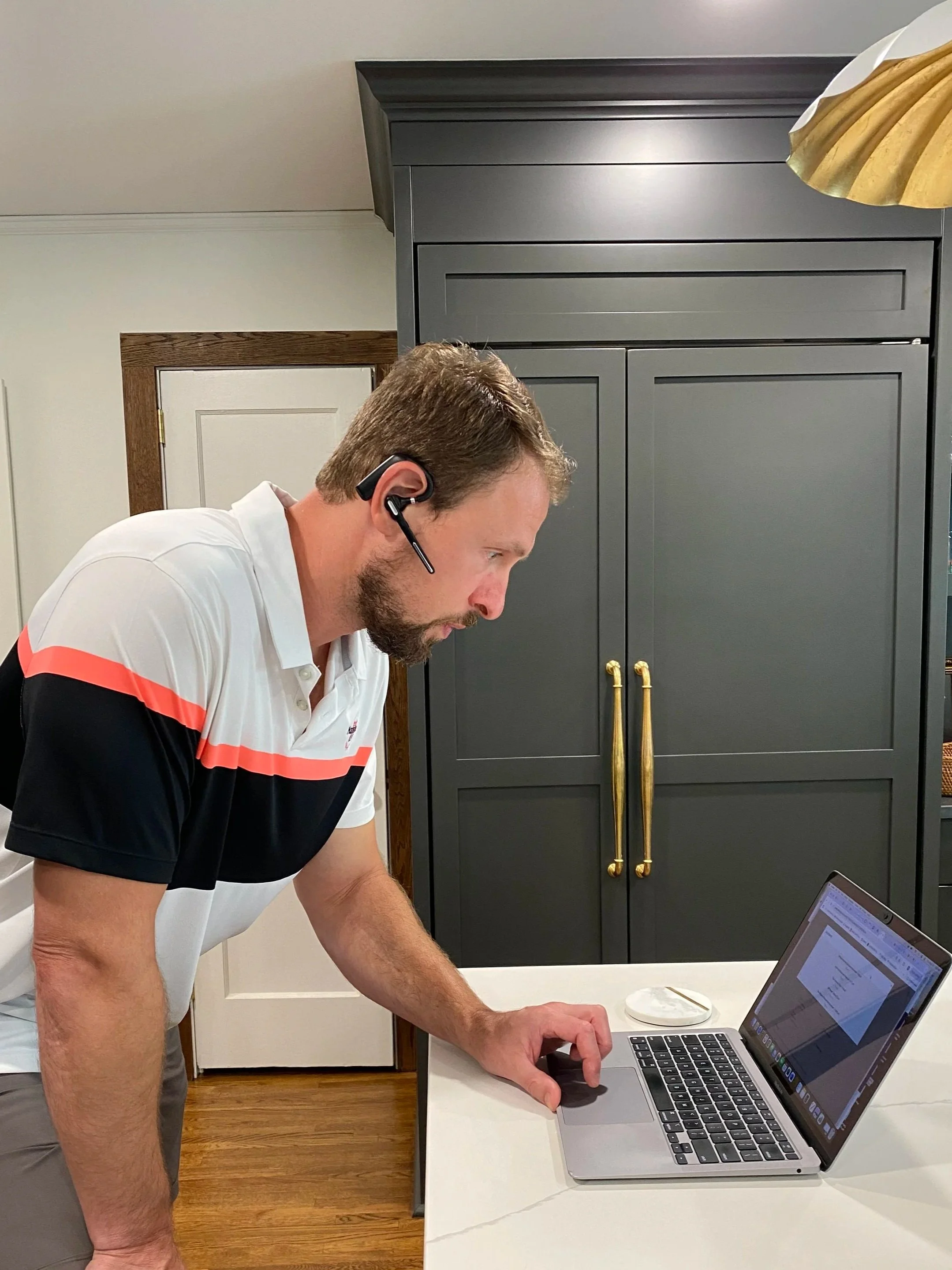 A man working at a desk with a laptop, wearing a headset, in a room with a dark cabinet and a door in the background.