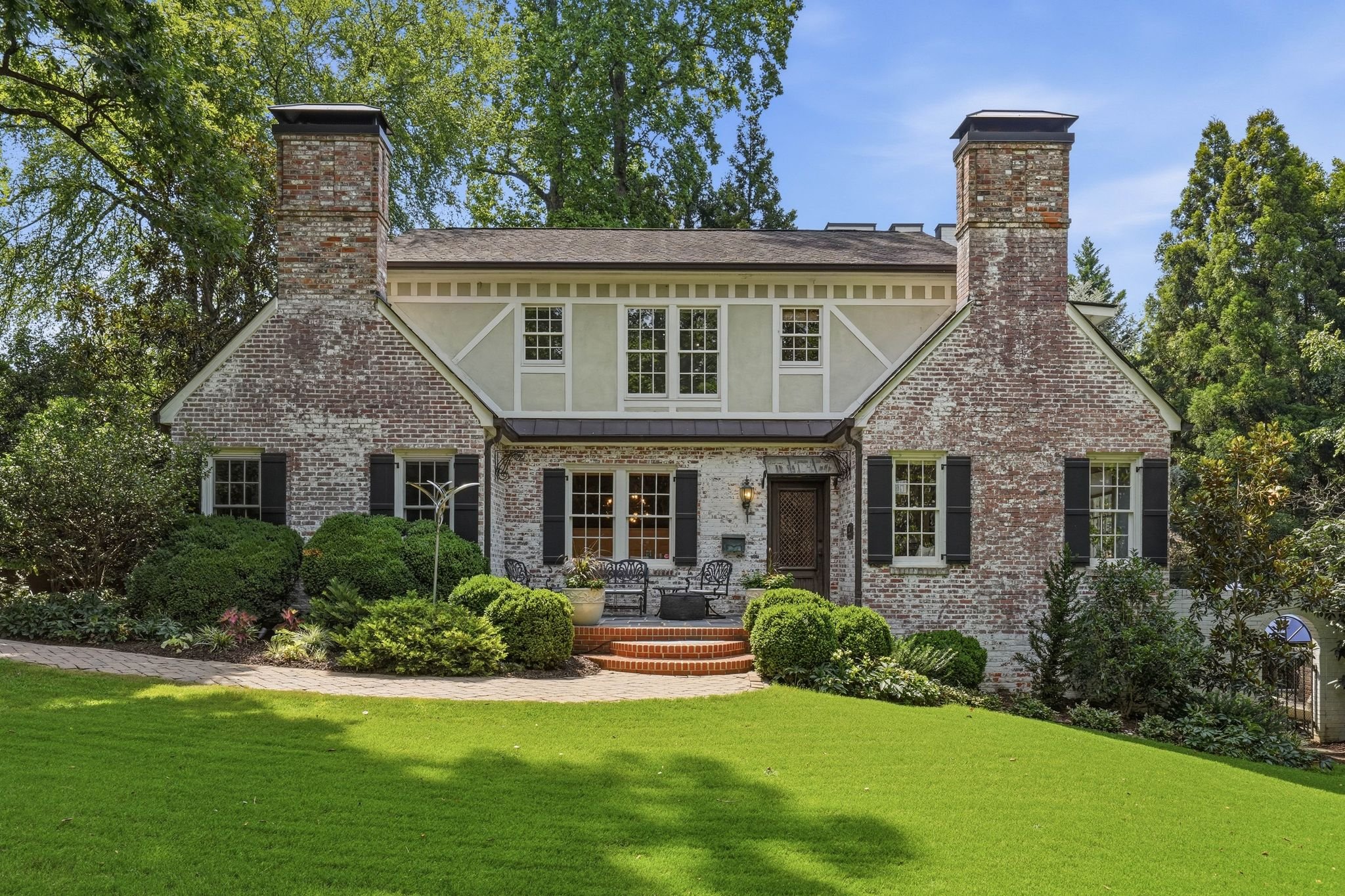 A two-story house in Plaza Midwood with a brick exterior, white trim, black shutters, and two prominent brick chimneys, surrounded by a lush green lawn and mature trees under a clear blue sky.