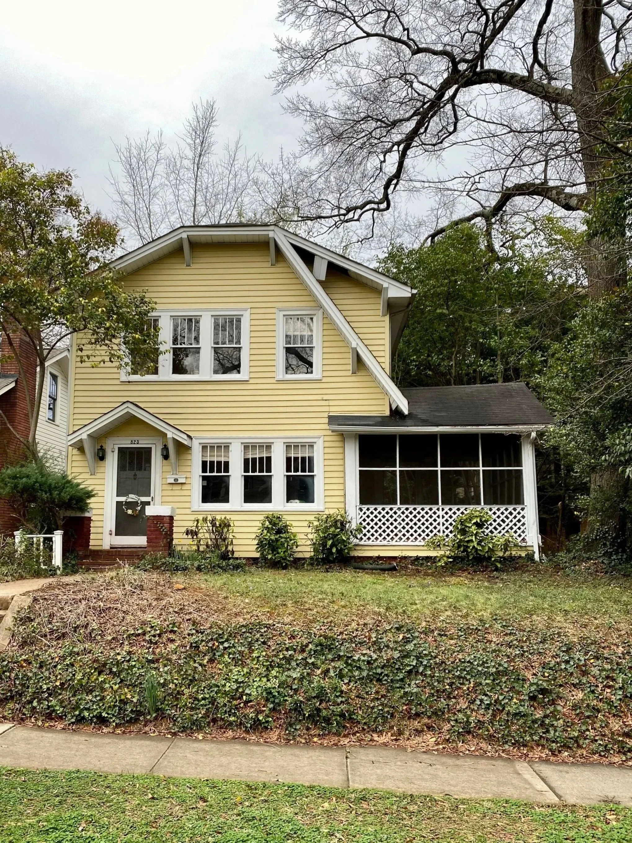 A yellow house with white trim and a screened porch, surrounded by trees and a garden, under an overcast sky.