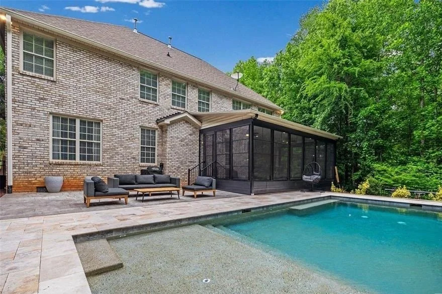 Backyard view of a large brick house with a swimming pool, outdoor seating area, and screened porch, surrounded by green trees under a blue sky.