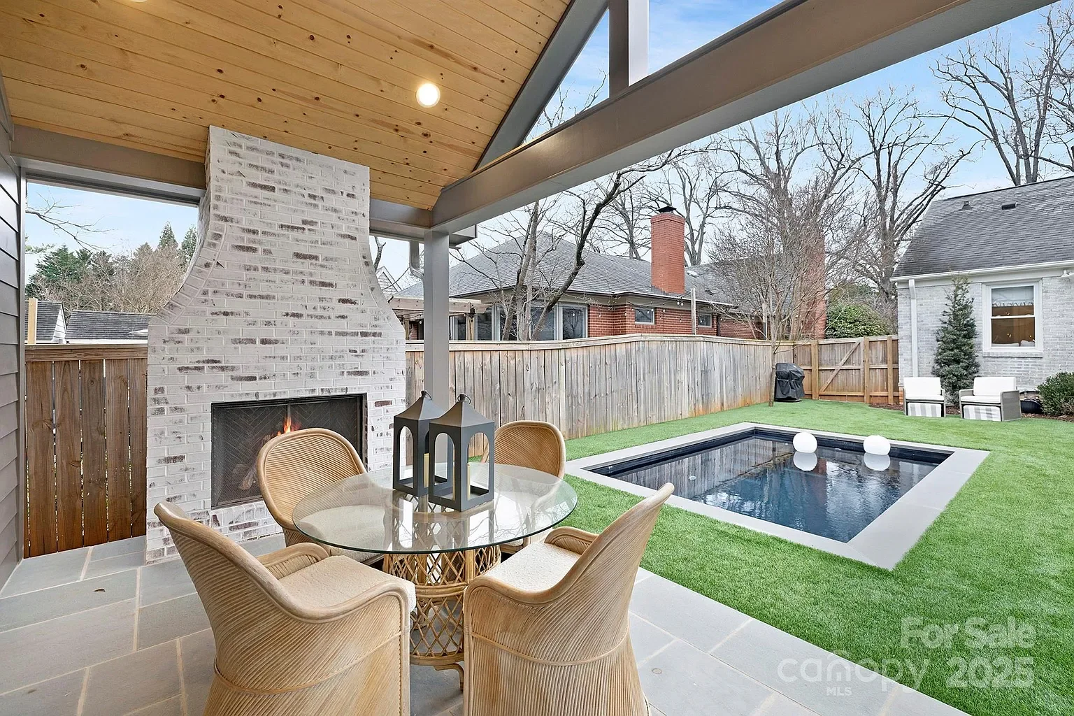 Covered backyard patio with a brick fireplace, dining table with chairs, and a small rectangular pool with floating spheres, surrounded by a wooden fence and outdoor seating