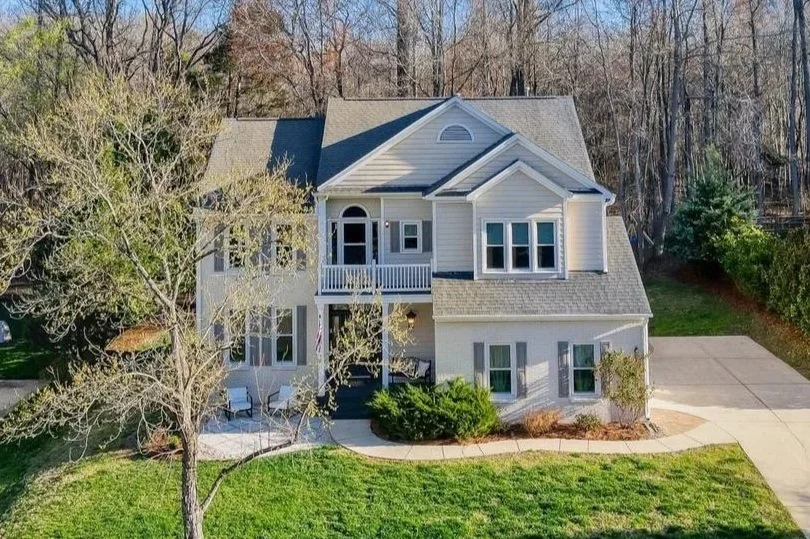 A two-story house with a gray roof, cream-colored siding, and multiple windows, surrounded by trees and a green lawn.