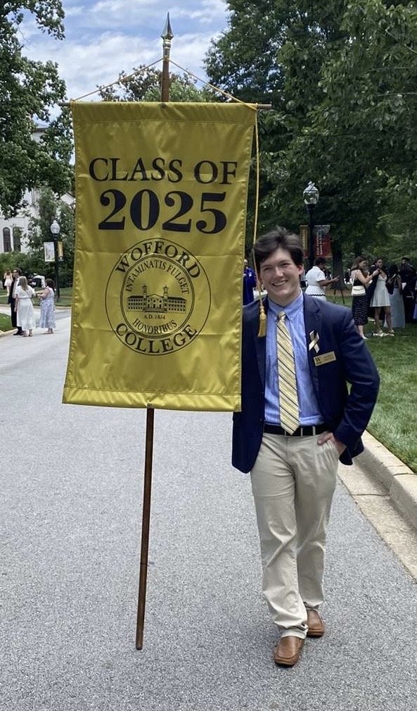 A young man dressed in a blue blazer, light-colored trousers, and a striped tie standing outdoors, holding a yellow banner that reads "Class of 2025" and features the emblem of Wooford College.