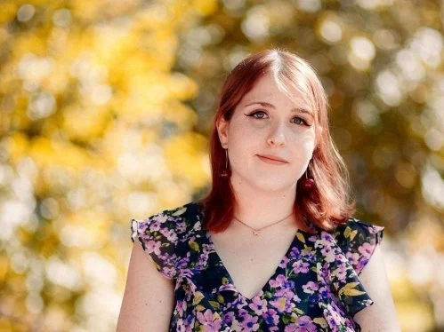 Young woman with red hair wearing a floral dress outdoors in front of fall-colored trees.