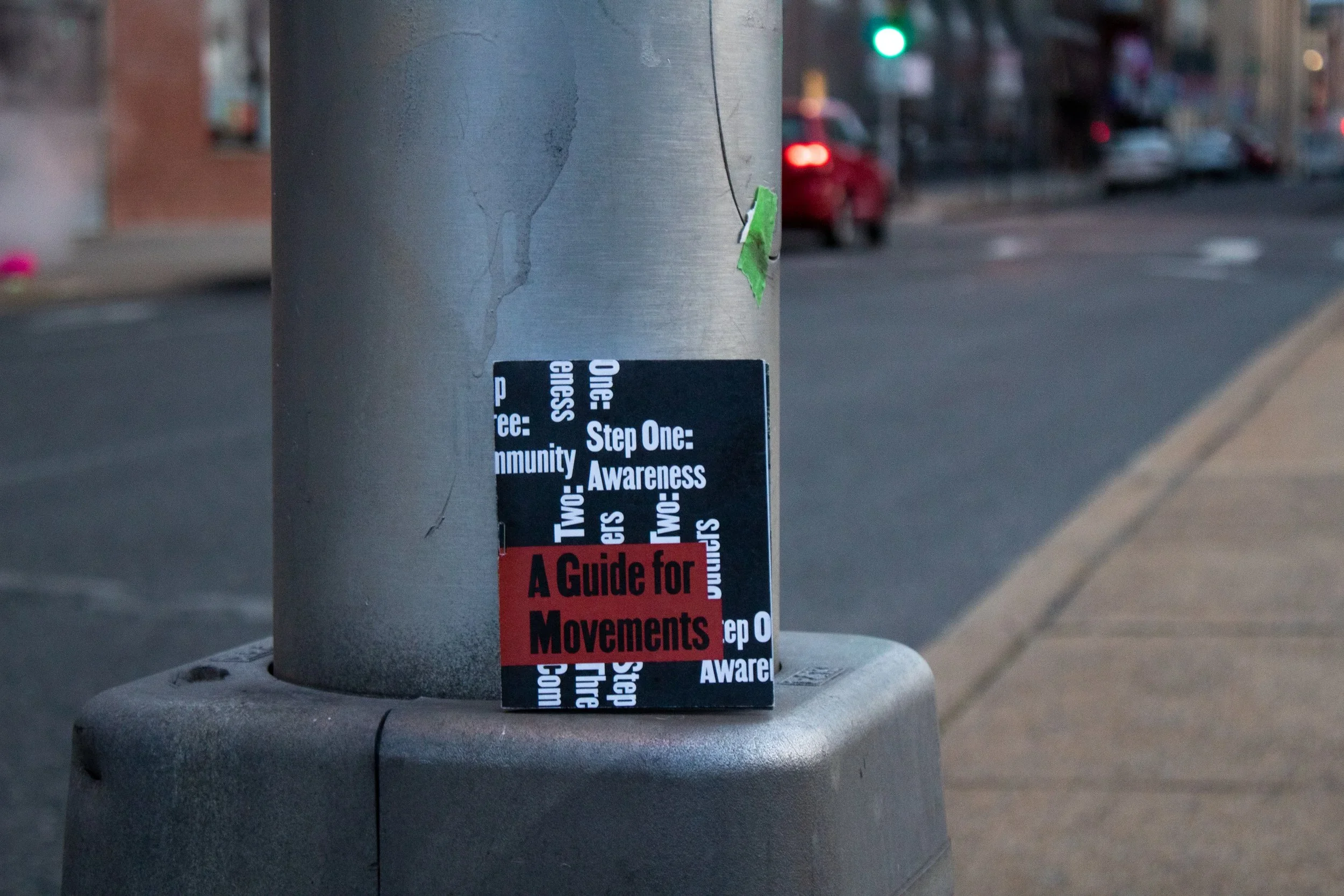 A black and red booklet titled 'A Guide for Movements' is attached to a gray street pole, with a sticker featuring words like 'One,' 'Step One,' 'Awareness,' and 'Community' partially visible.