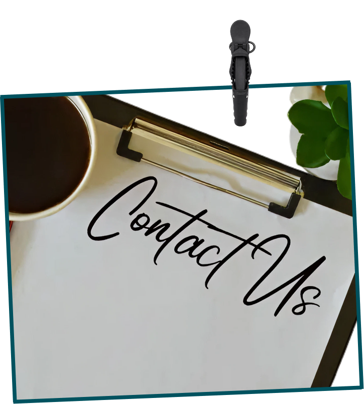 A clipboard with a sign that reads 'Contact Us' in bold script font, a cup of coffee, a green plant, and a portion of a white mug around the clipboard on a workspace surface.