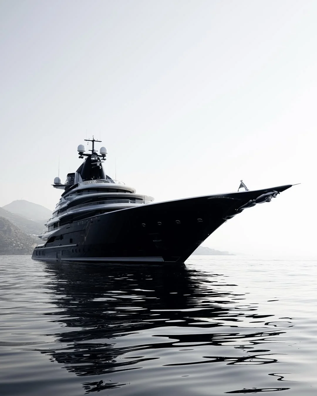A large, black luxury yacht floating on calm water, with a faint outline of hills in the background and a clear, bright sky.