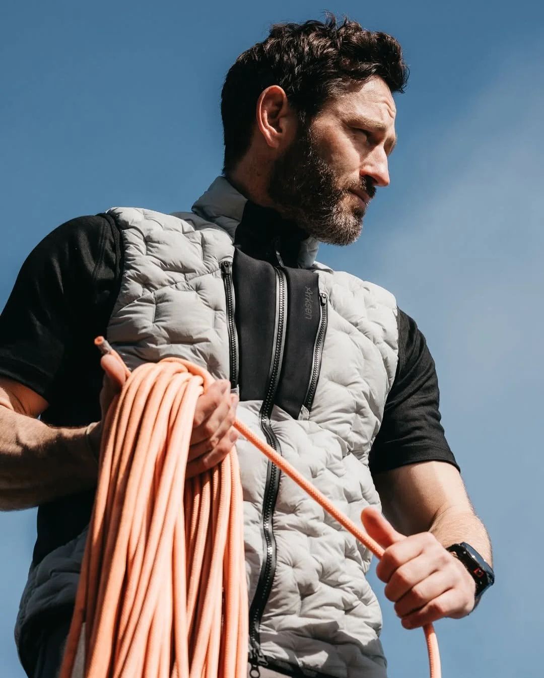 A man with a beard and dark hair, dressed in outdoor gear, holding climbing rope against a clear blue sky.