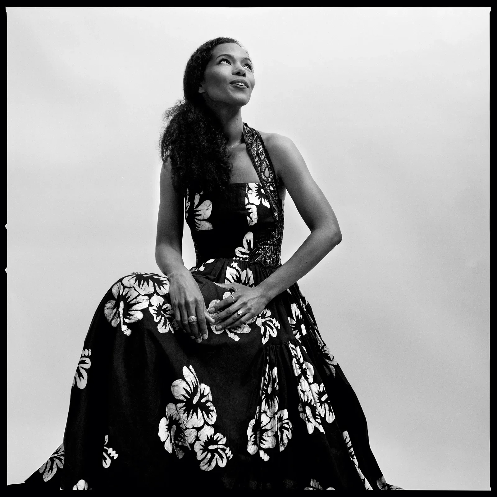 A woman in a floral dress, sitting on a bench, looking up in black and white photograph.