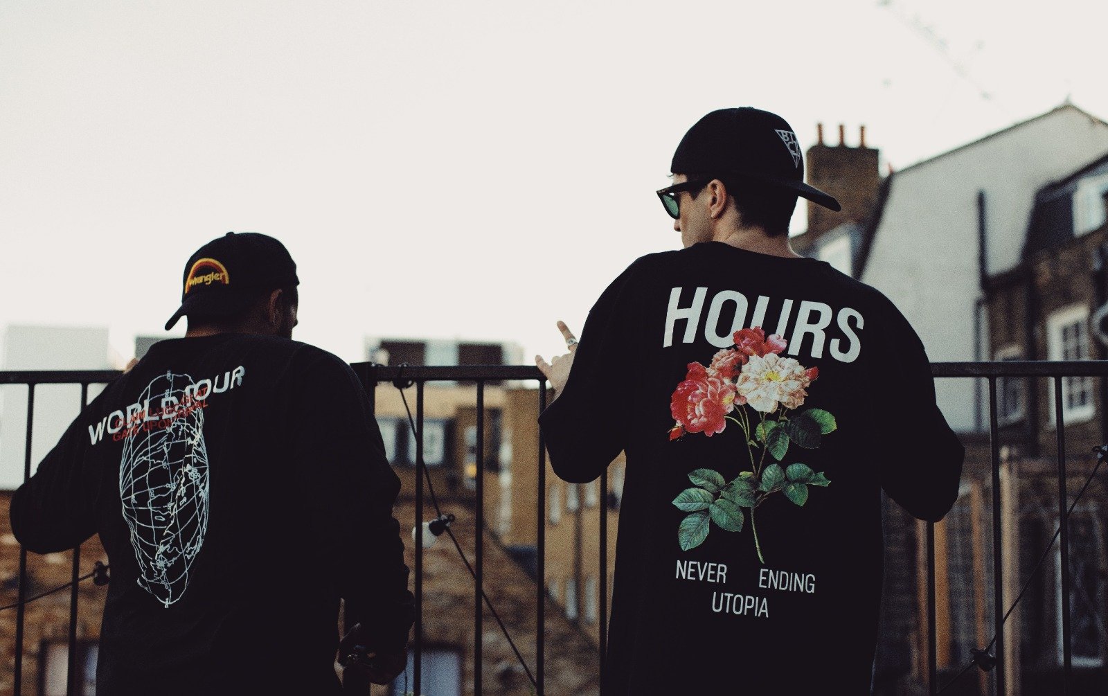 Two young men wearing black hoodies and caps, standing by a metal fence in an urban area, with buildings in the background. One hoodie has a floral design with the text "NEVER ENDING UTOPIA," and the other has graphics and text related to "WORLD AGENDA."
