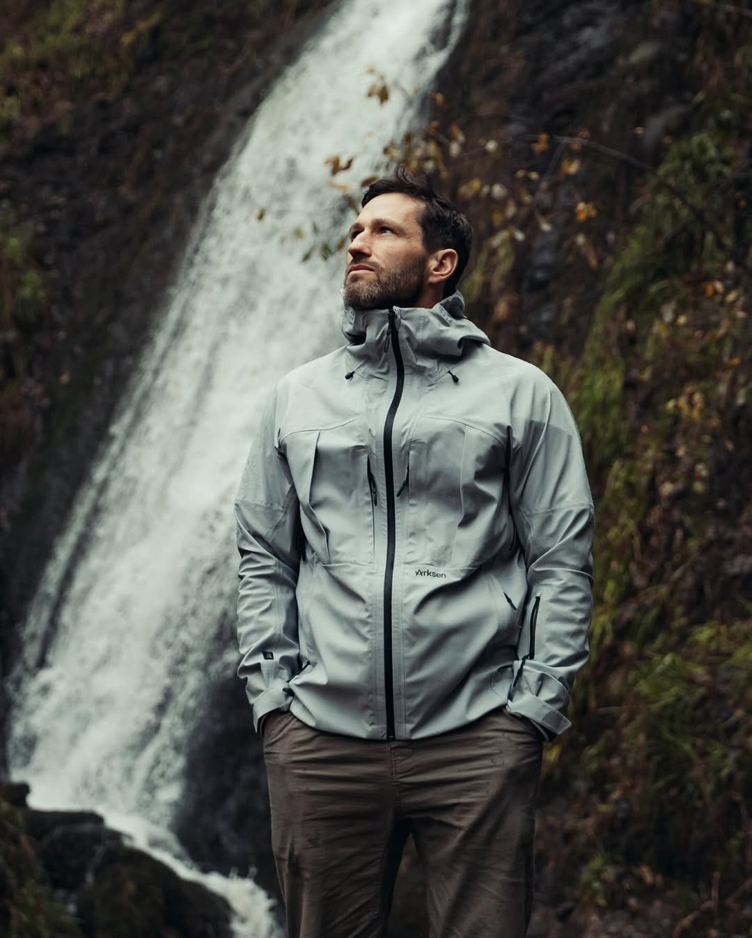 A man in outdoor gear standing near a waterfall with a contemplative expression.