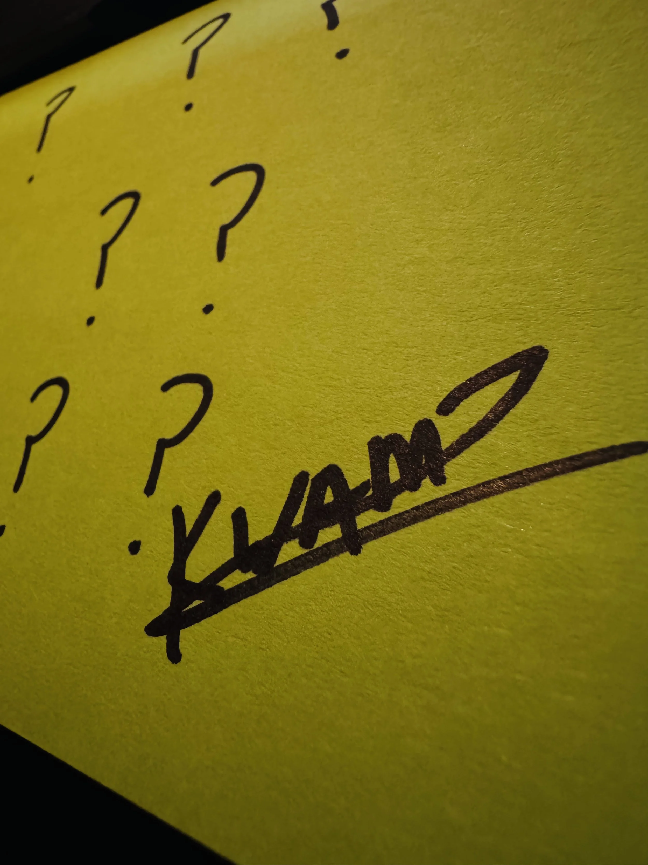 Yellow paper with multiple black question marks and the name 'KLAUS' written in black ink, with a line crossing out the name.