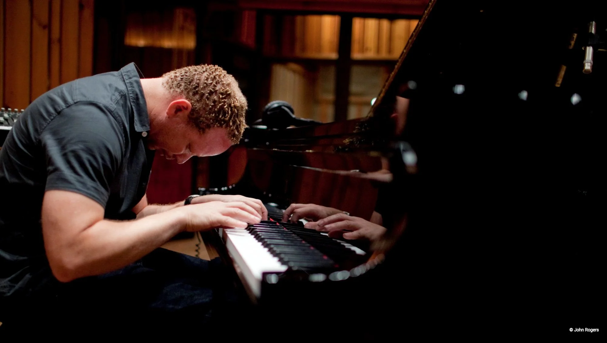 A person with curly short hair, wearing a black shirt, leaning over a grand piano with closed eyes, playing the keys in a wooden recording studio or rehearsal space.