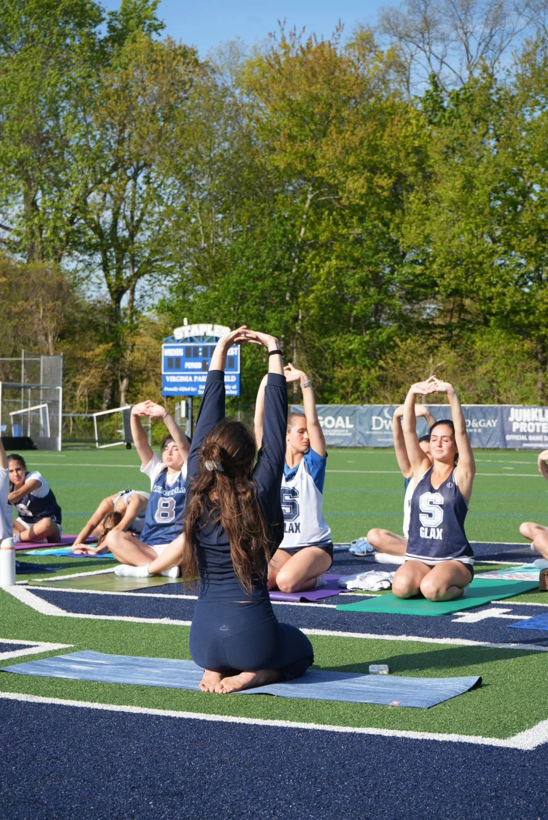 Young women participating in an outdoor yoga class on a sports field, sitting cross-legged on yoga mats, stretching with arms raised overhead on a sunny day with trees in the background.