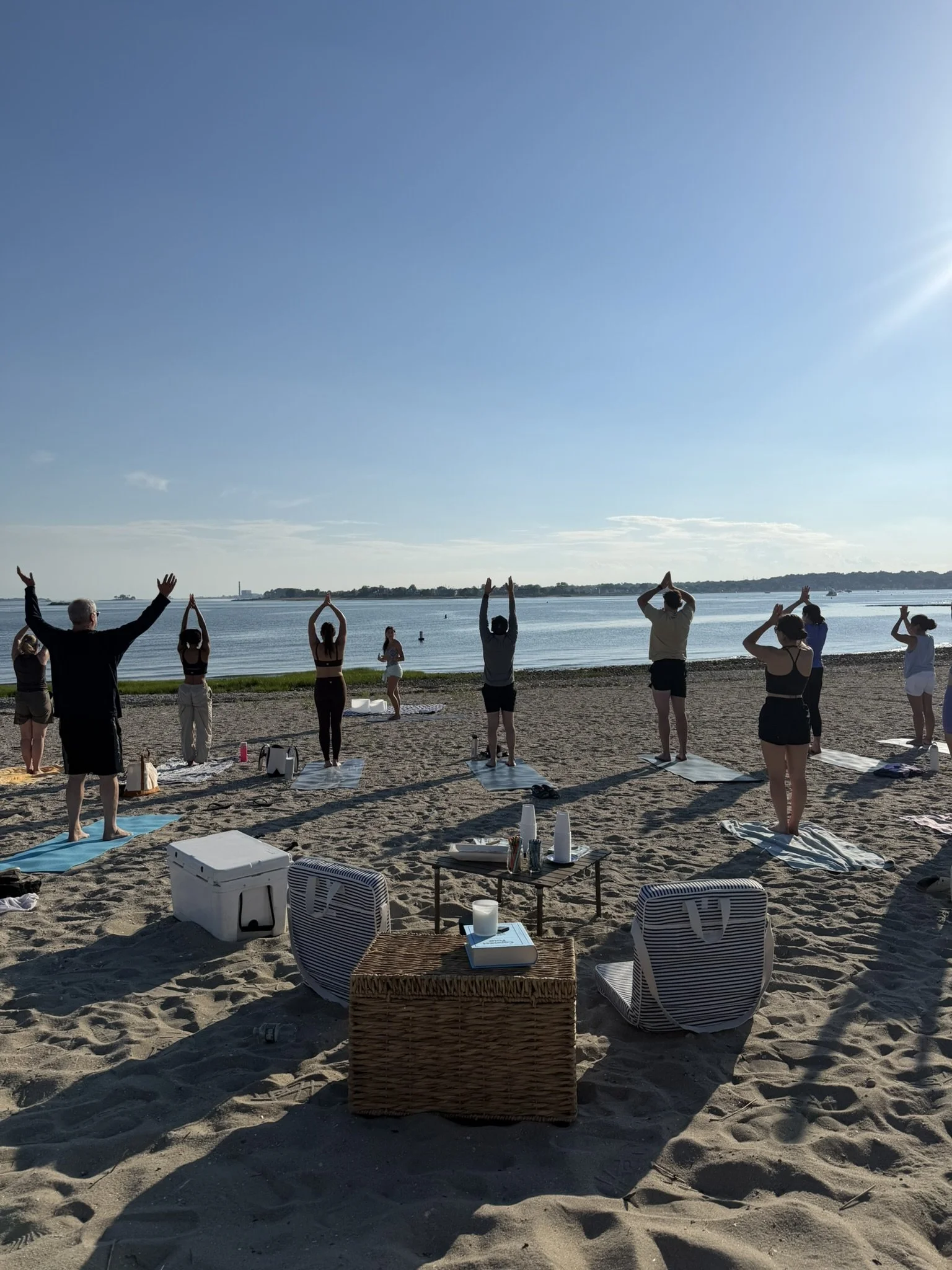 Group of people practicing yoga on the beach near a body of water, with some standing on mats with their arms raised or in prayer poses as the sun shines brightly overhead.