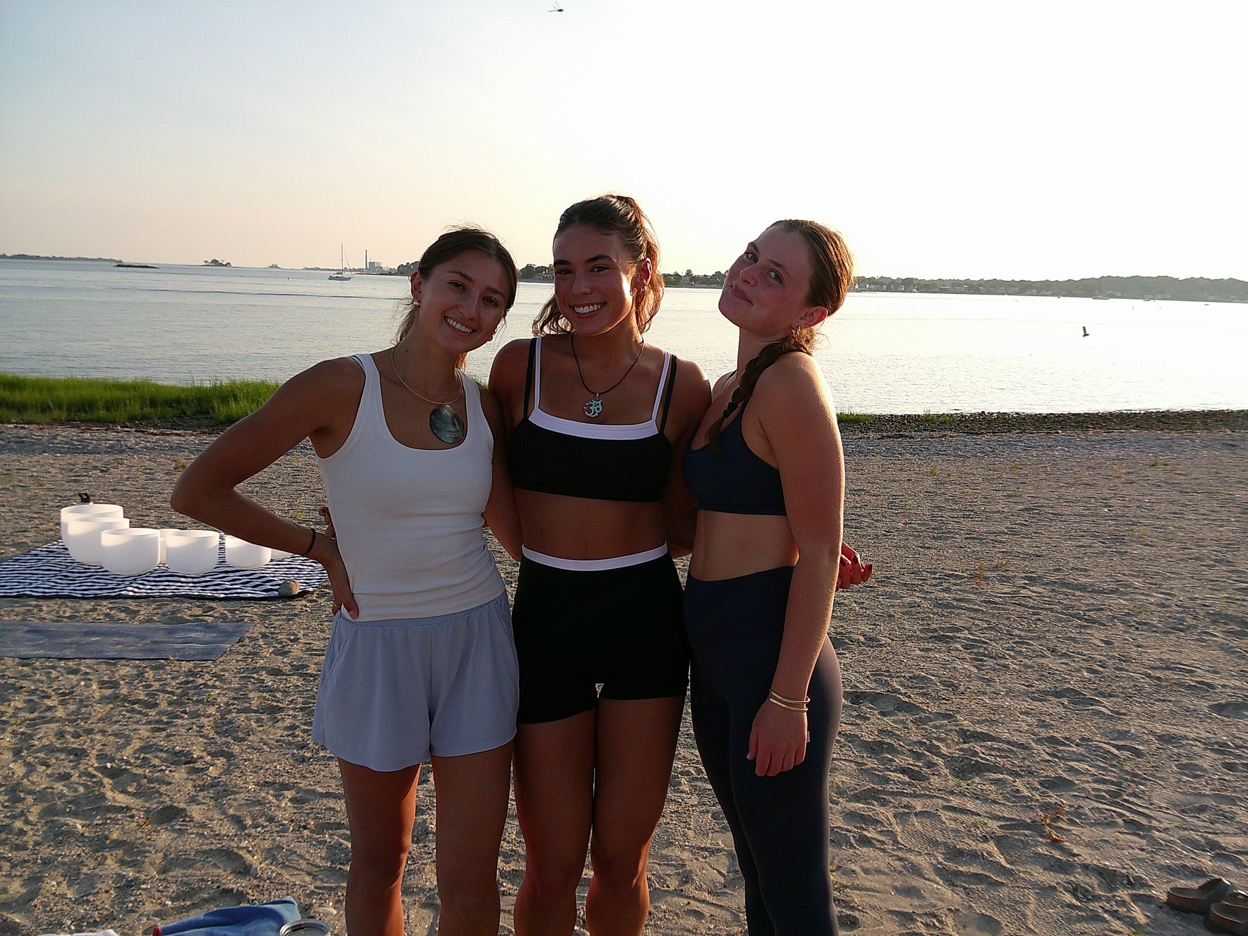 Three women standing close together on a beach during sunset, with water and sailboats in the background. They are smiling and wearing casual summer clothes.