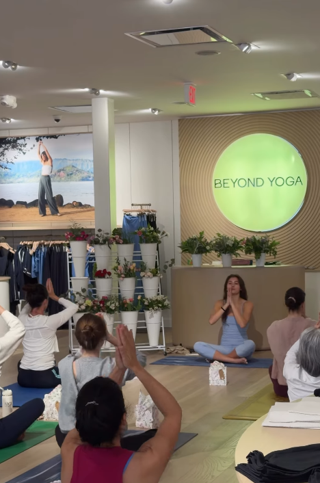 People attending a yoga class in a studio with a instructor sitting cross-legged in front, leading the session. A screen displays 'BEYOND YOGA.'