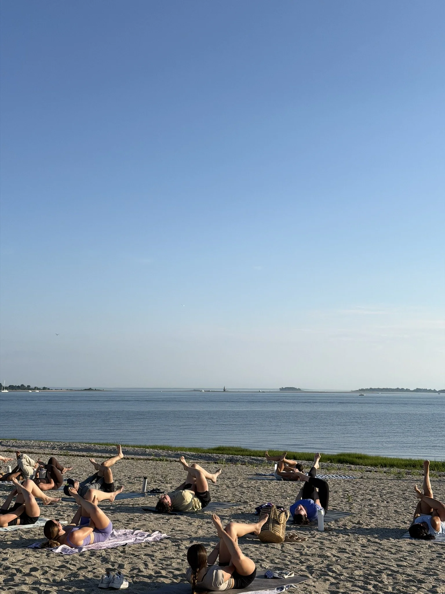 People practicing yoga on a sandy beach by the water during daytime, with a clear blue sky and distant boats on the water.