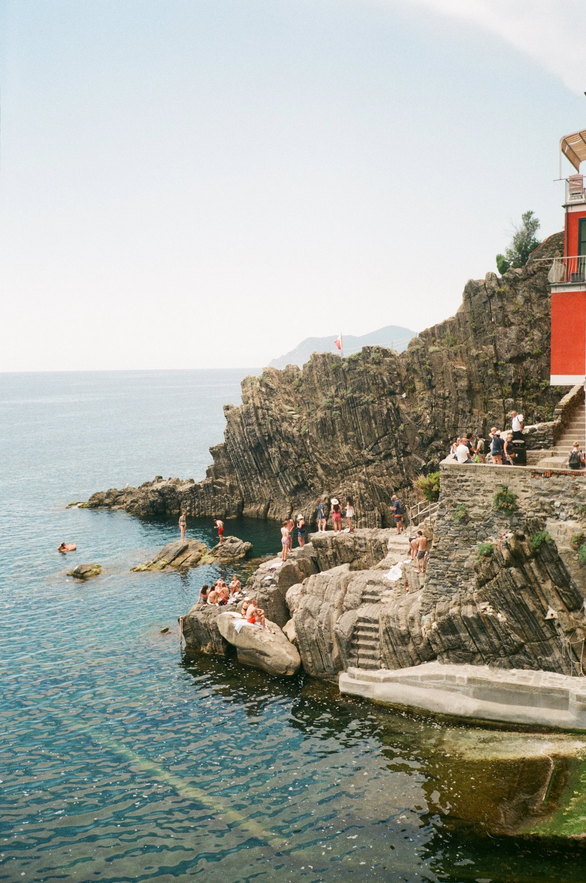People relaxing and swimming near rocky seaside with cliffs, building, and flag in the background.