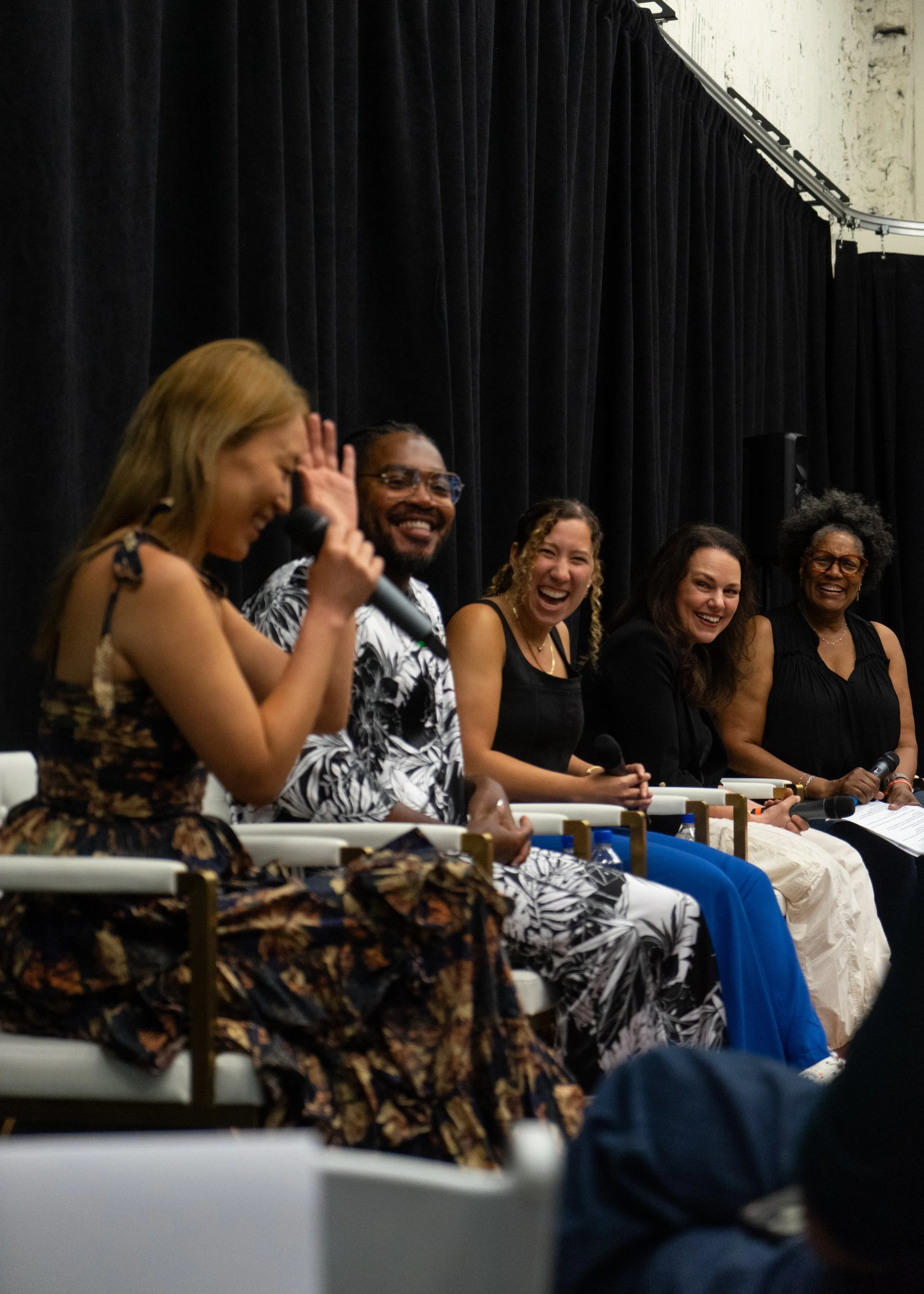 A group of five women sitting at a panel discussion or interview, smiling and laughing. They are seated in front of black curtains and holding microphones.