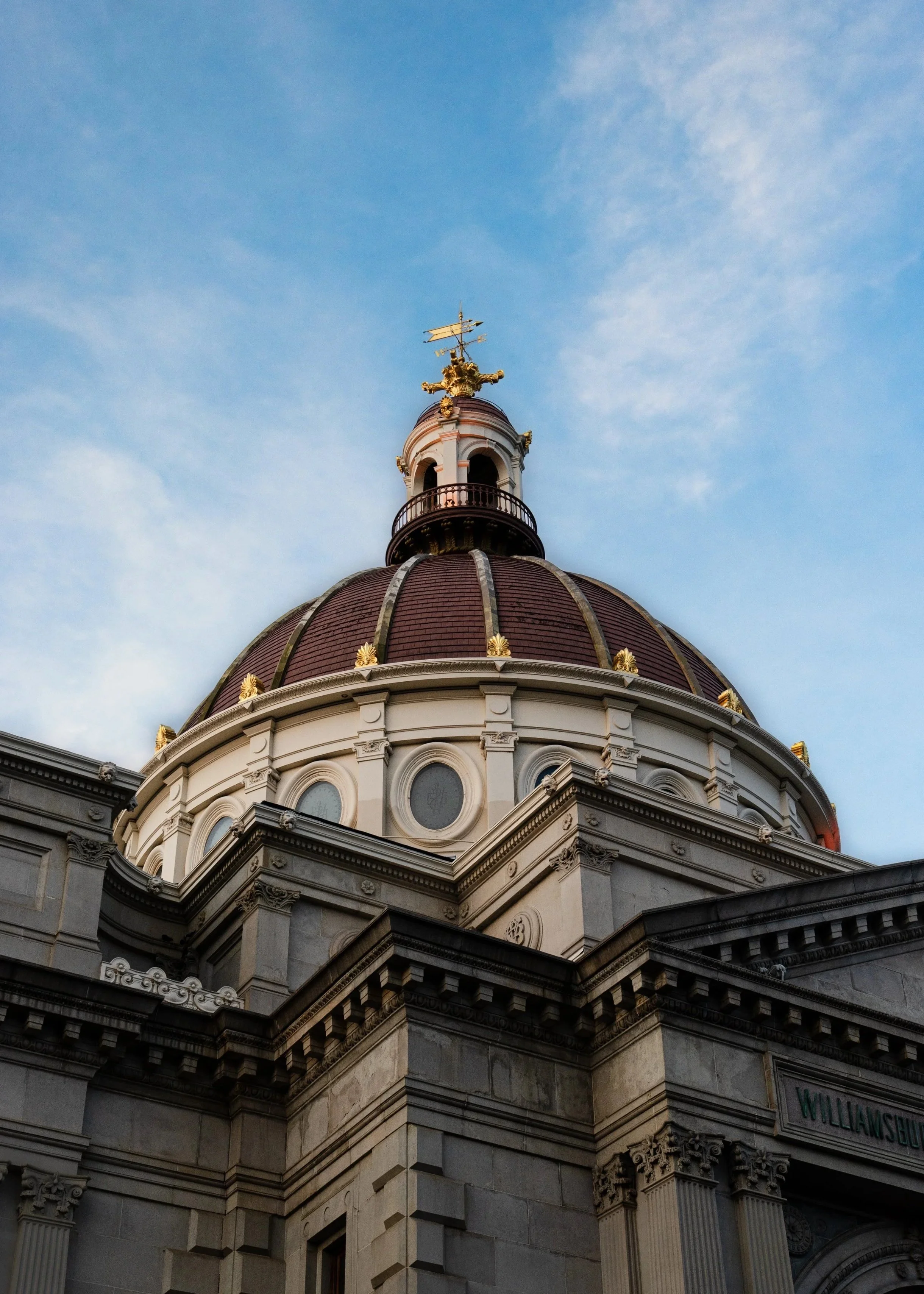 Close-up view of a historic building dome with ornate architecture, gold accents, and a weather vane on top, against a blue sky with scattered clouds.