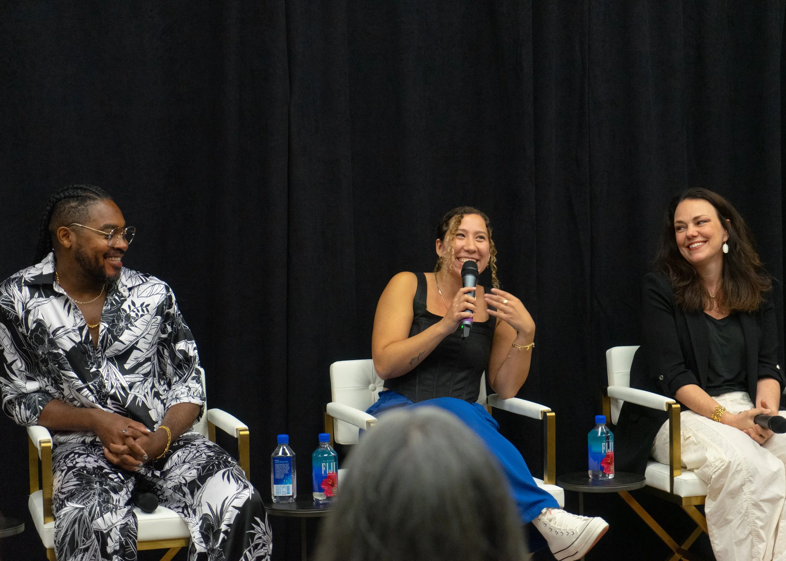 Three people sitting on a panel at an event, two women and one man, with black curtains in the background. The woman in the middle is holding a microphone and speaking, smiling. Bottled water is on the small table in front of them.