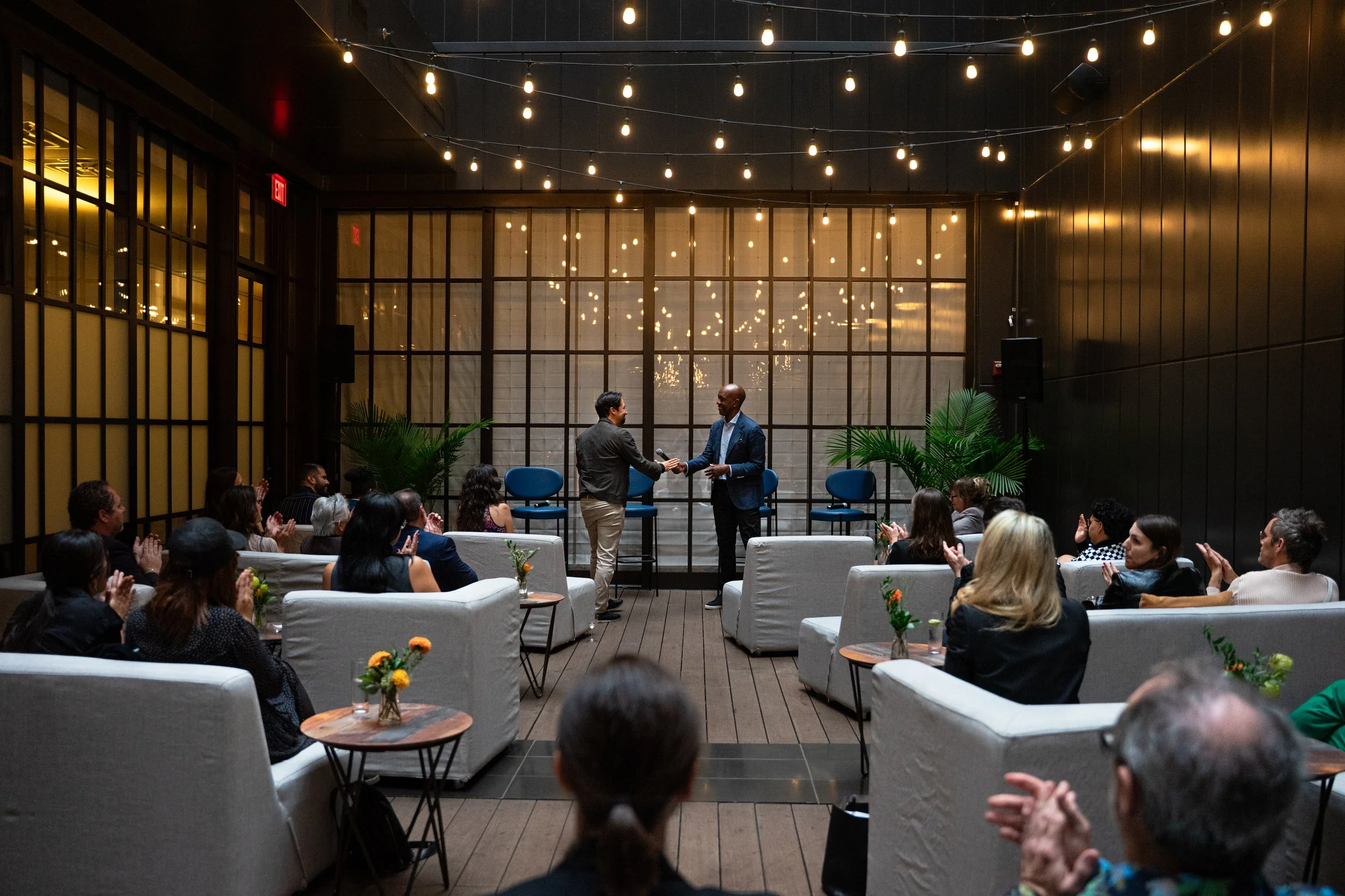 Two men shaking hands on stage in a dimly lit event space with string lights overhead, audience seated on sofas and chairs clapping, plants and small tables with flowers in the foreground.