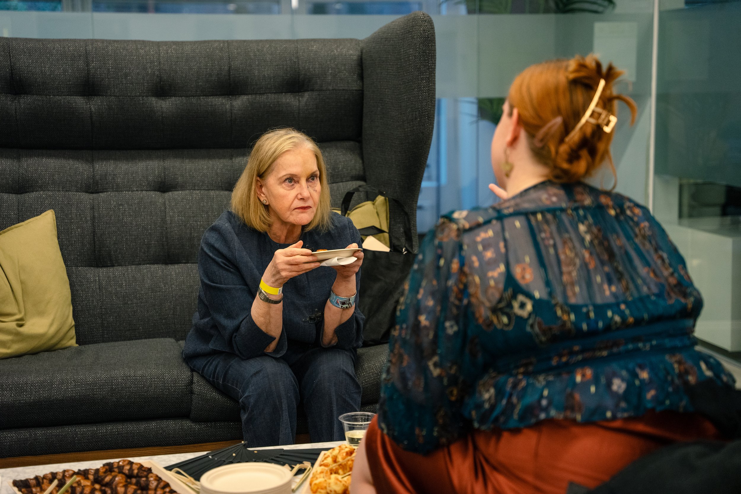 Two women having a conversation at a social gathering with food on a table in front of them. One woman has blonde hair and is holding a small plate, the other woman has red hair in a bun with a hair clip and is wearing a patterned top.