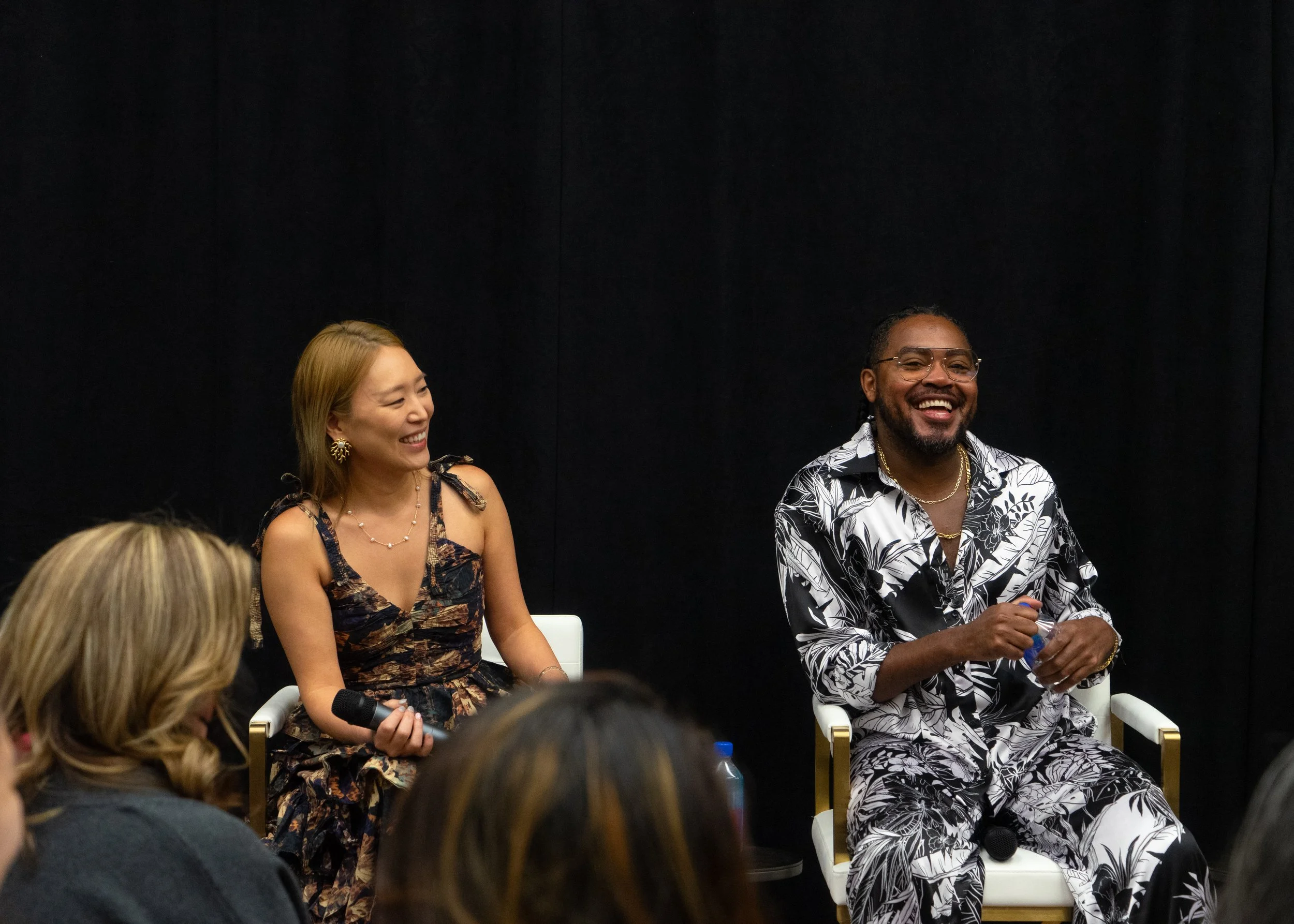 Two people sitting and smiling at a panel discussion or event. The woman on the left has long hair, is dressed in a floral dress, and is holding a microphone. The man on the right has glasses, a beard, and is dressed in a patterned shirt, holding a w