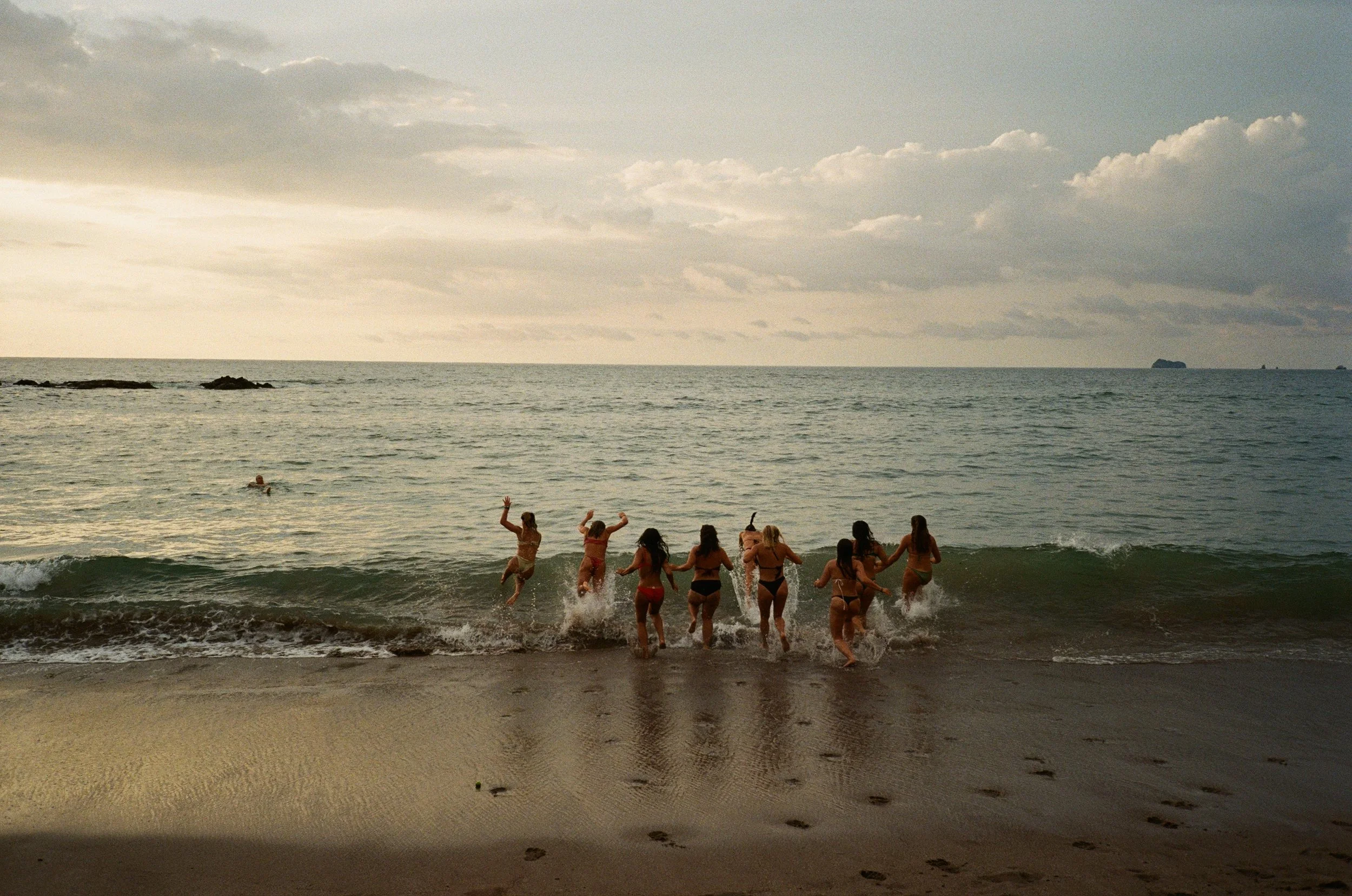 Group of women in bikinis playing and running in the ocean waves at sunset or dusk, with rocky outcroppings and islands in the distance under a cloudy sky.