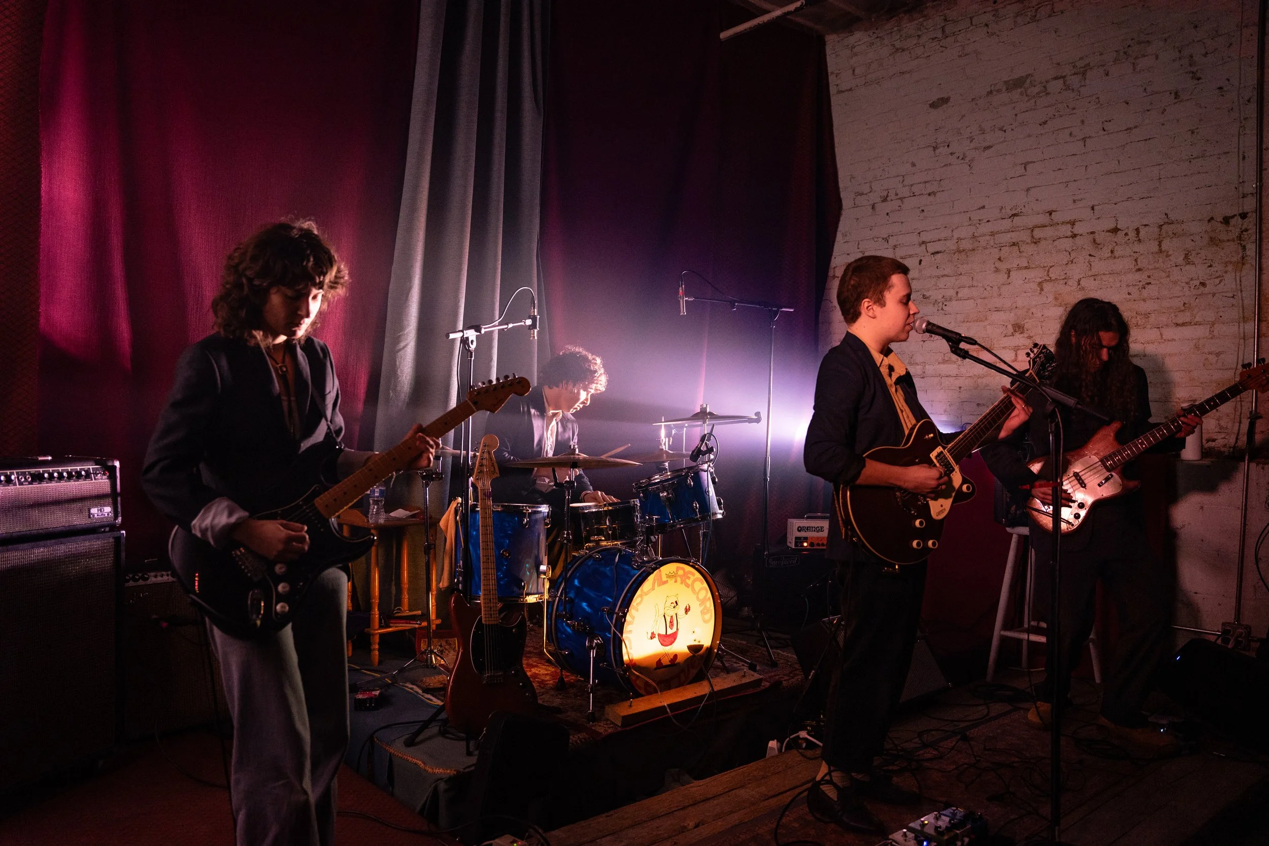 A band performing on stage with four musicians playing instruments, illuminated by stage lighting and set against a brick wall backdrop.