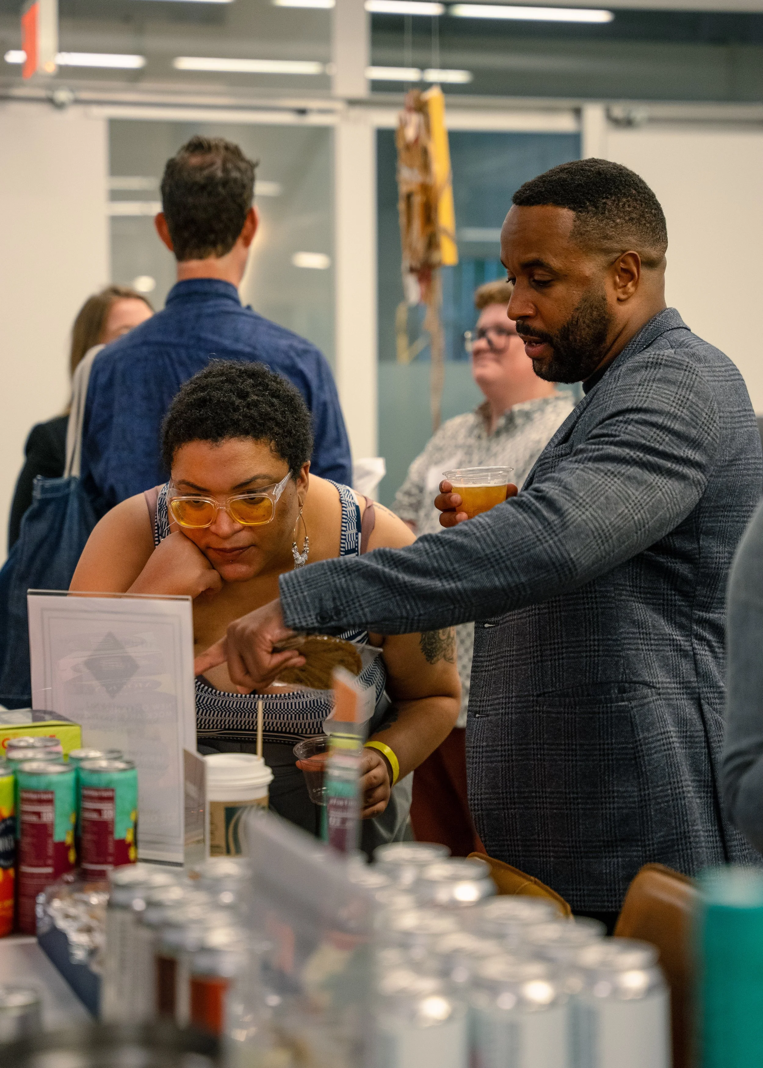 People gathered at a booth or table at an indoor event or market, with various canned drinks and food items displayed in the foreground. One woman is wearing glasses and a sleeveless top, looking down at her phone or a tablet. A man beside her, dress