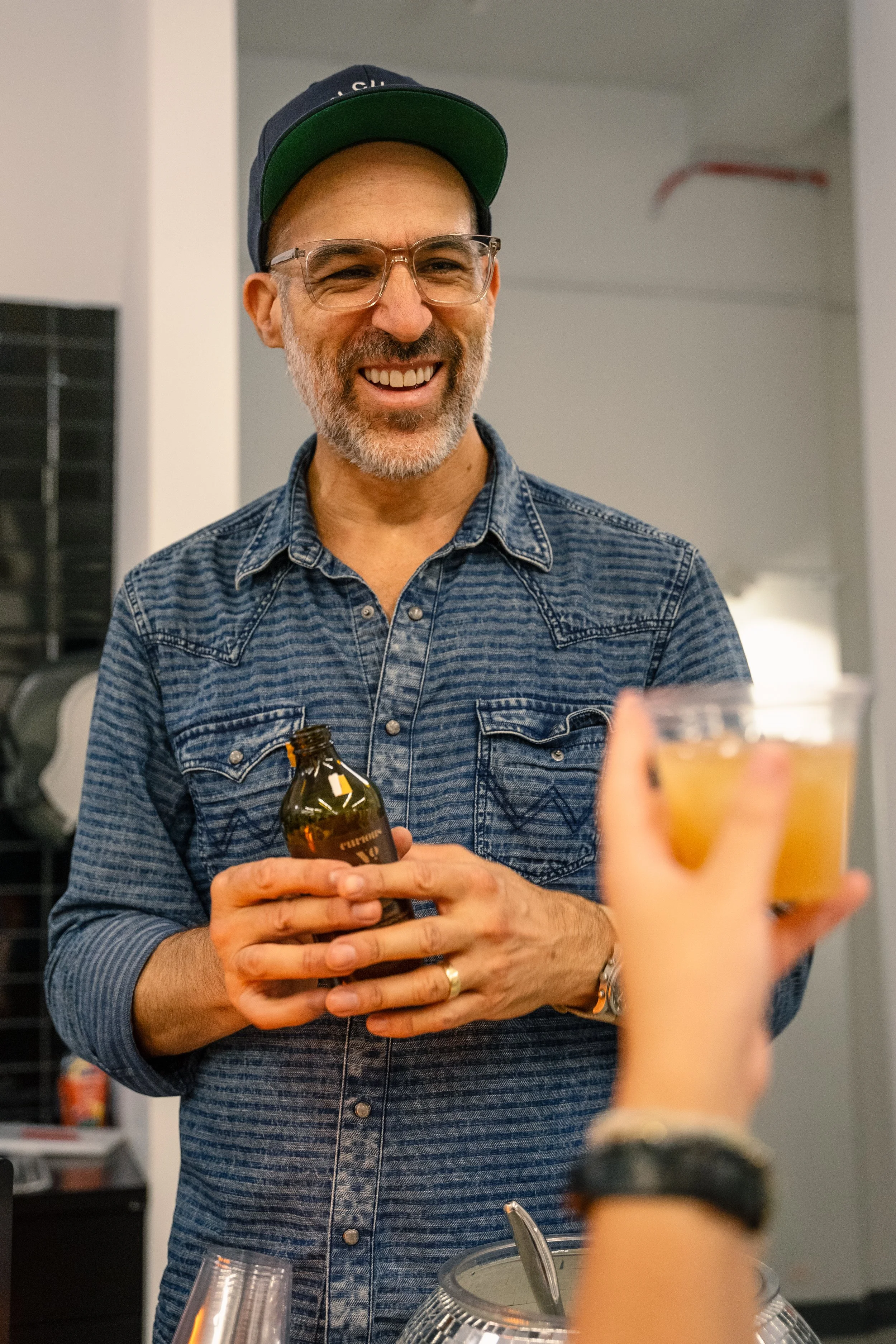 A man with glasses and a beard wearing a denim shirt and a cap, smiling and holding a beer bottle, at a social gathering.
