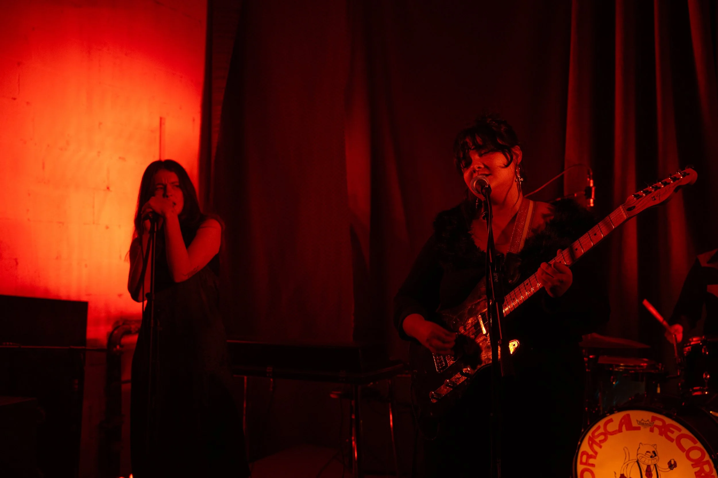 Two women performing on stage with red lighting. One is singing into a microphone, and the other is playing an electric guitar and singing into a microphone.