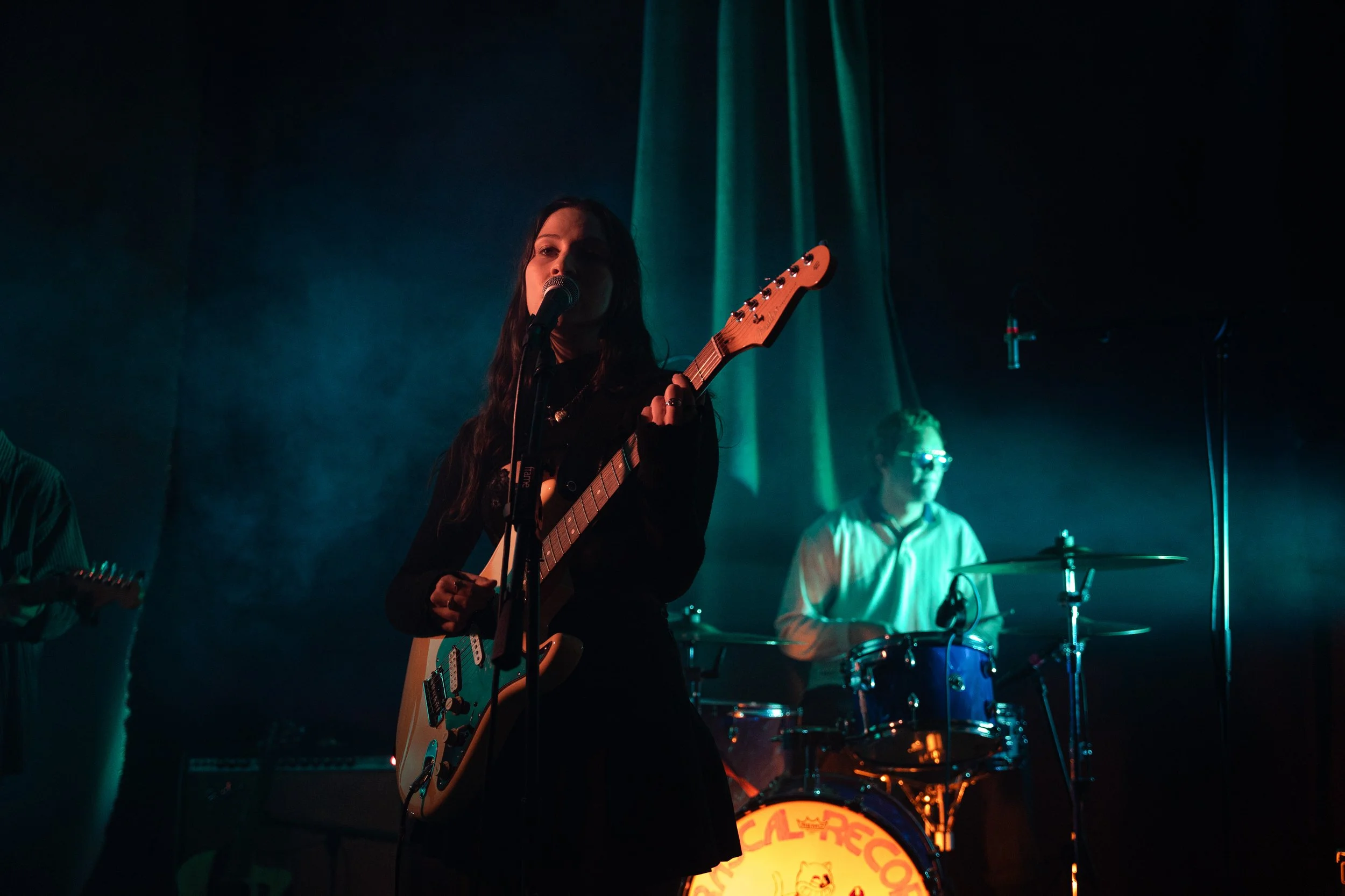 A woman with long dark hair playing an electric guitar and singing into a microphone on a dimly lit stage. Behind her, a man is playing drums. The stage has colored lighting, creating a vibrant musical atmosphere.