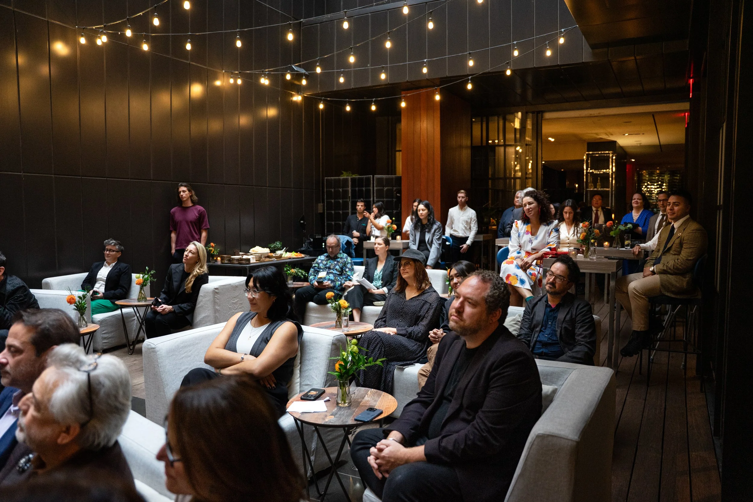 People gathered in a darkened indoor space with string lights hanging from the ceiling, sitting on couches and chairs, some taking notes and others listening attentively, indicating a social event or presentation.