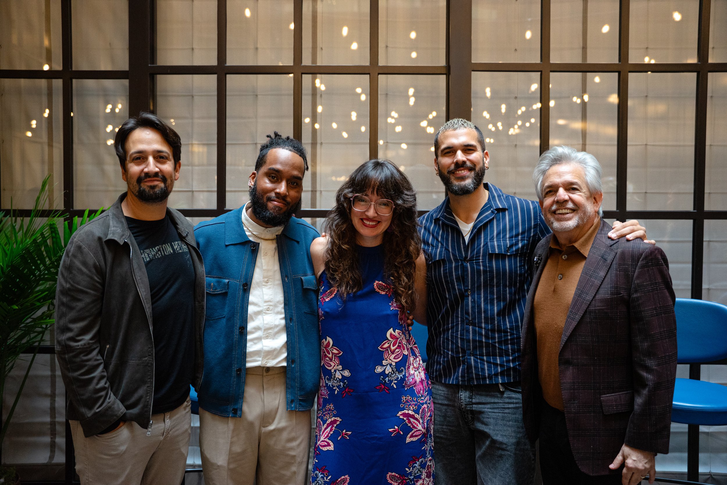 Group of five diverse people smiling and standing close together indoors in front of a large window with lights reflecting behind them.