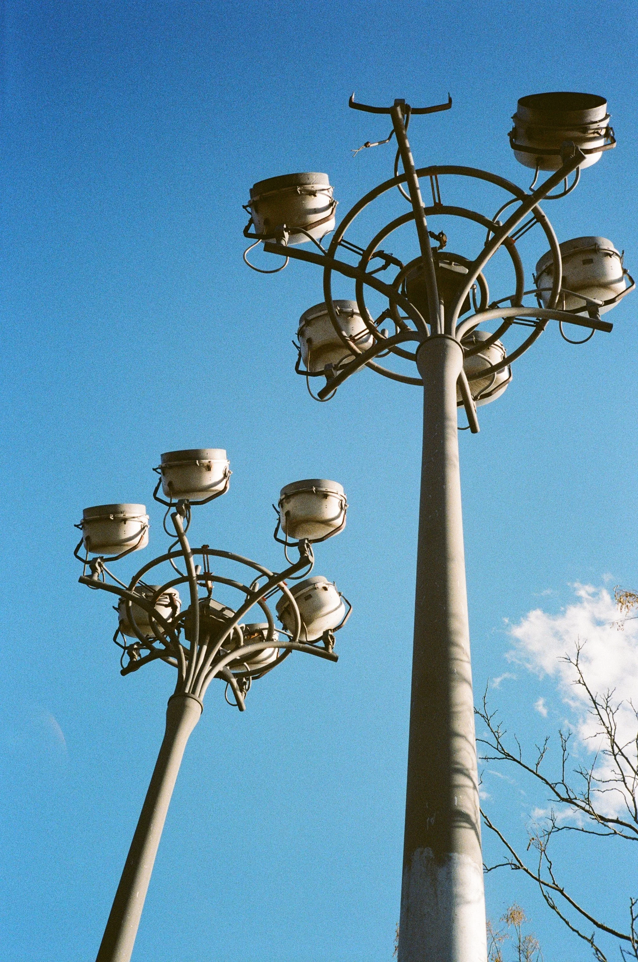 Two tall, modern streetlights with multiple circular lamp fixtures against a clear blue sky, with a few bare tree branches visible in the lower right.