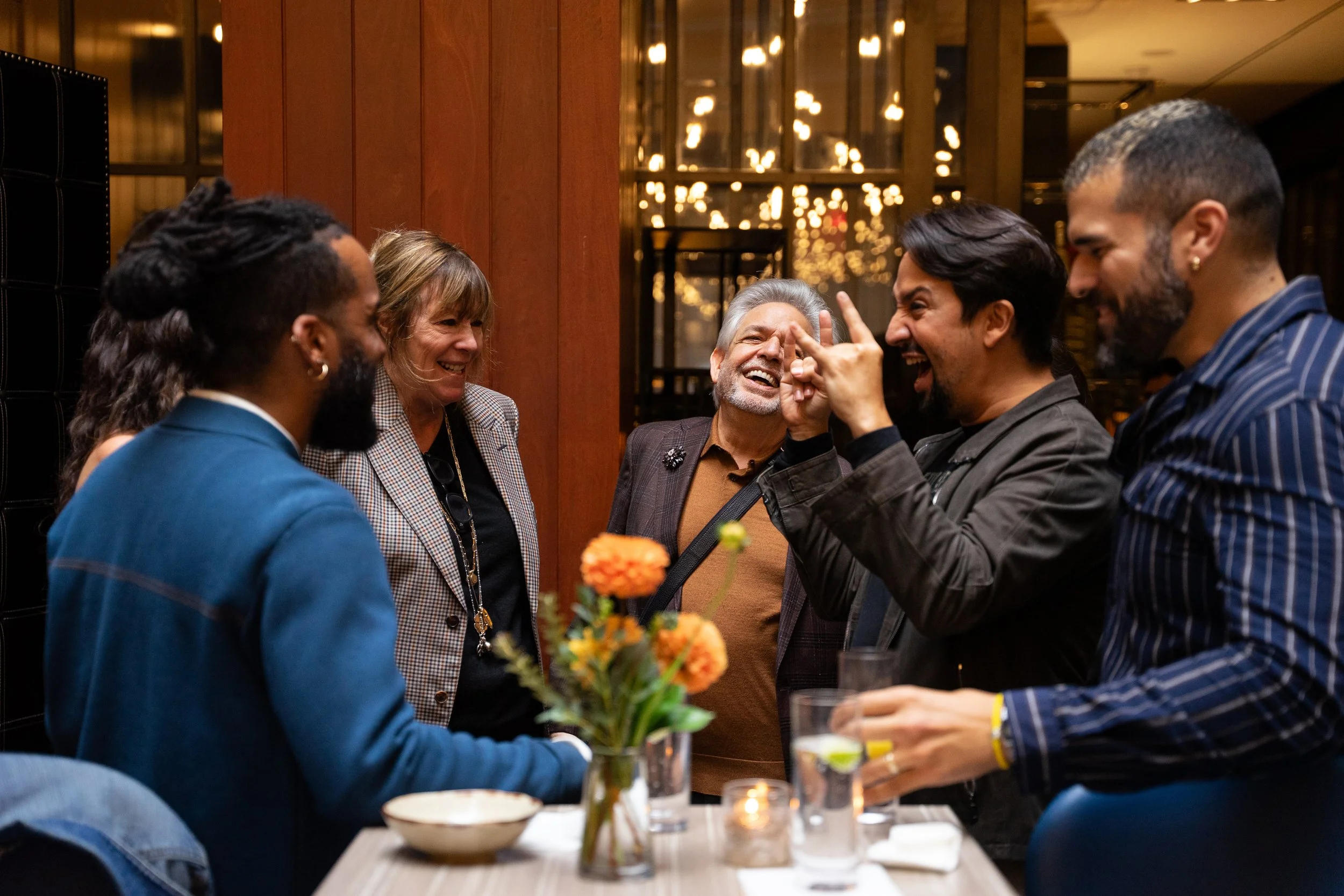 A group of five diverse adults gathered around a table, engaged in conversation and laughing, in a warmly lit restaurant or bar, with a floral centerpiece and drinks on the table.