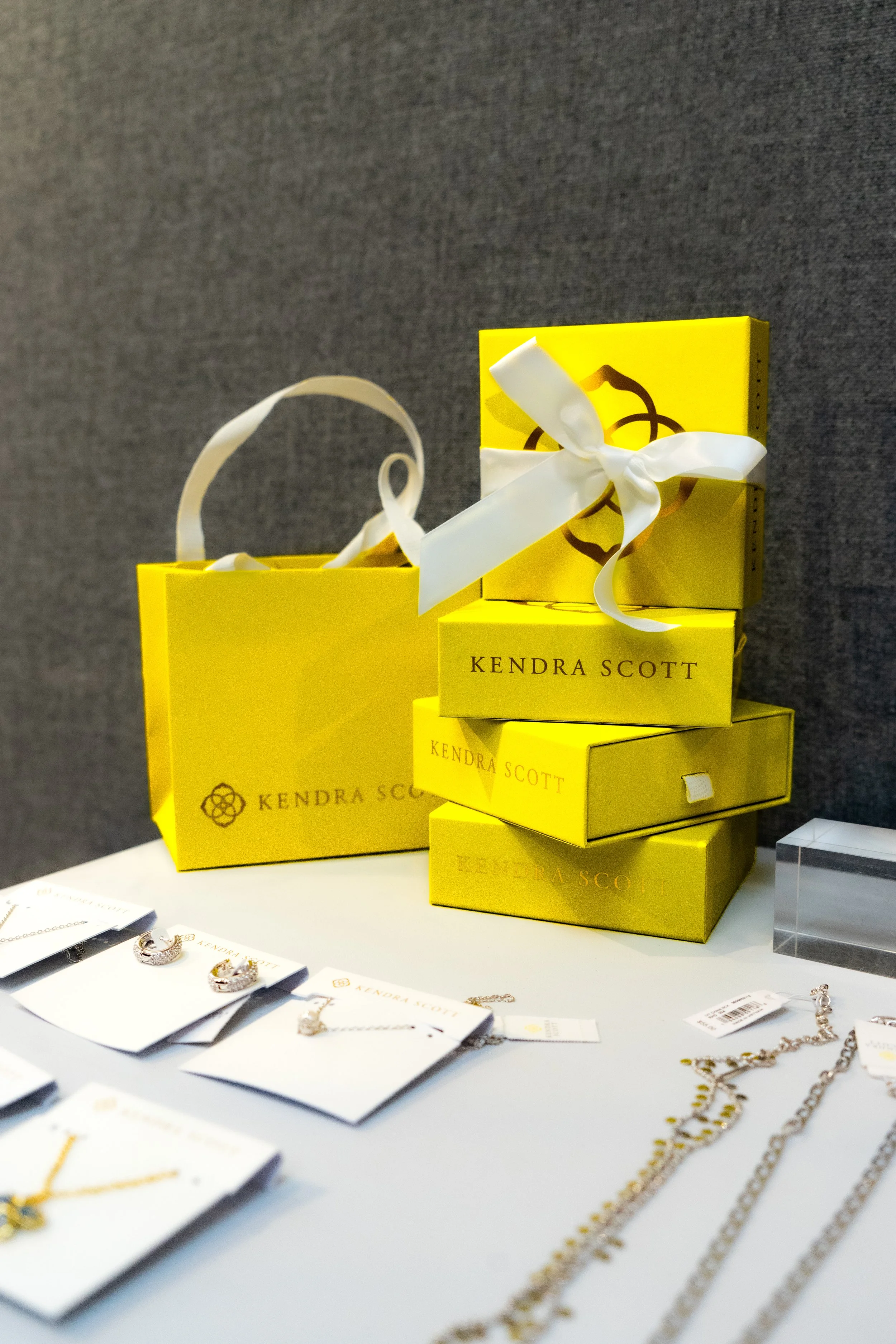 Display of jewelry including earrings and necklaces, with yellow boxes labeled 'KENDRA SCOTT' stacked in the background.