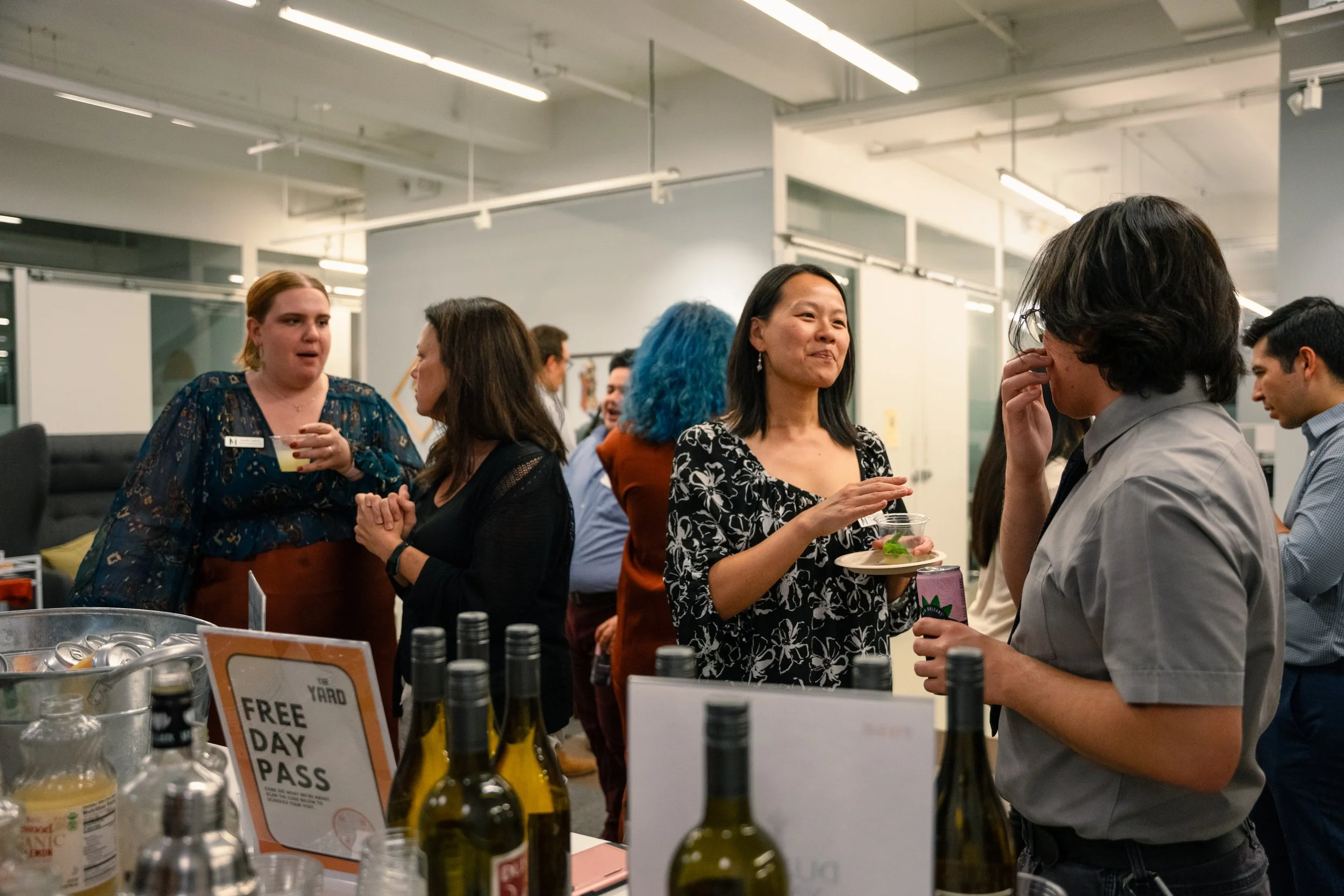People socializing at a booth with bottles and a sign that reads 'Free Day Pass' in an office or event space.
