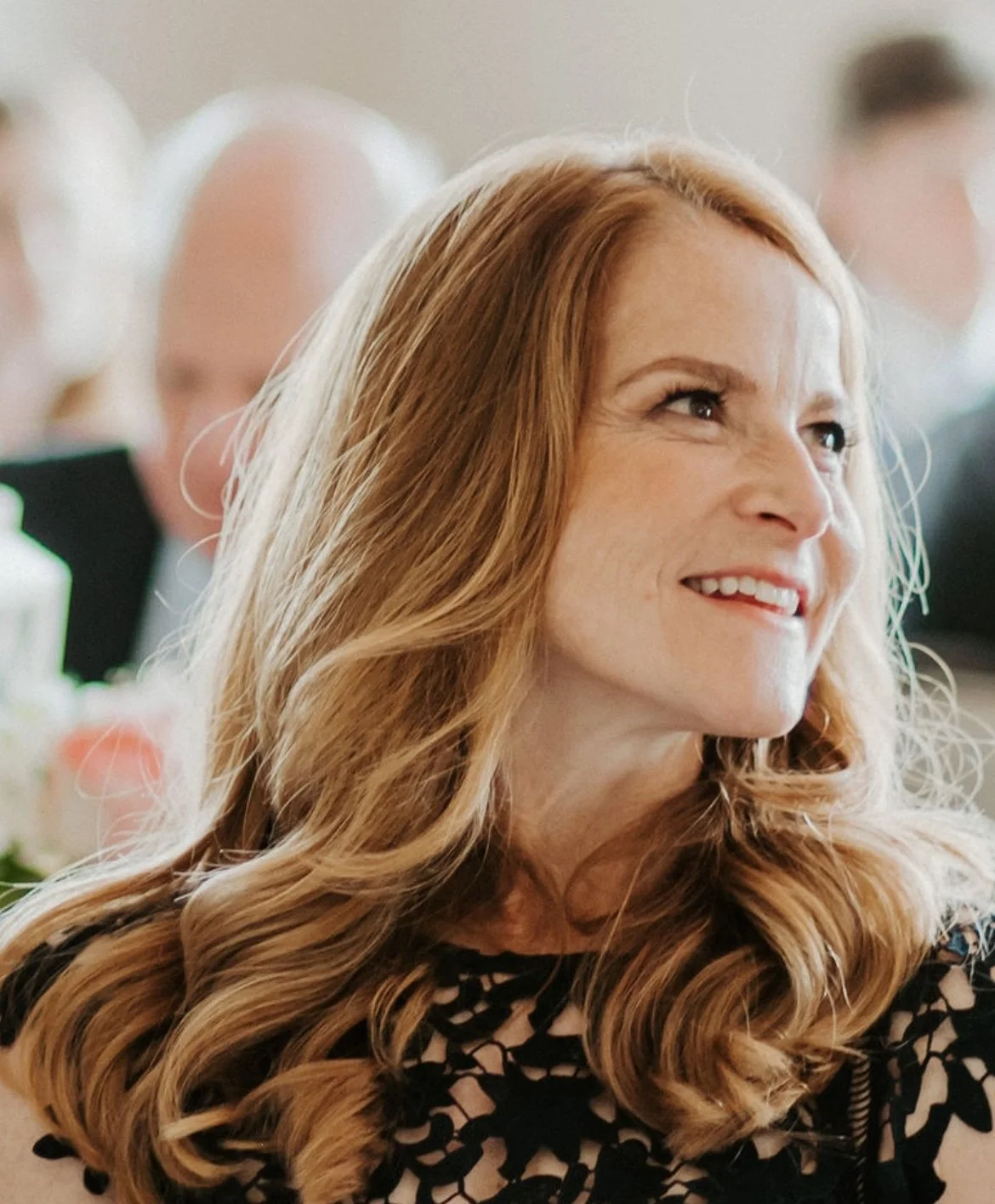 A smiling woman with long, wavy auburn hair wearing a black patterned dress, at a social event with blurred people in the background.