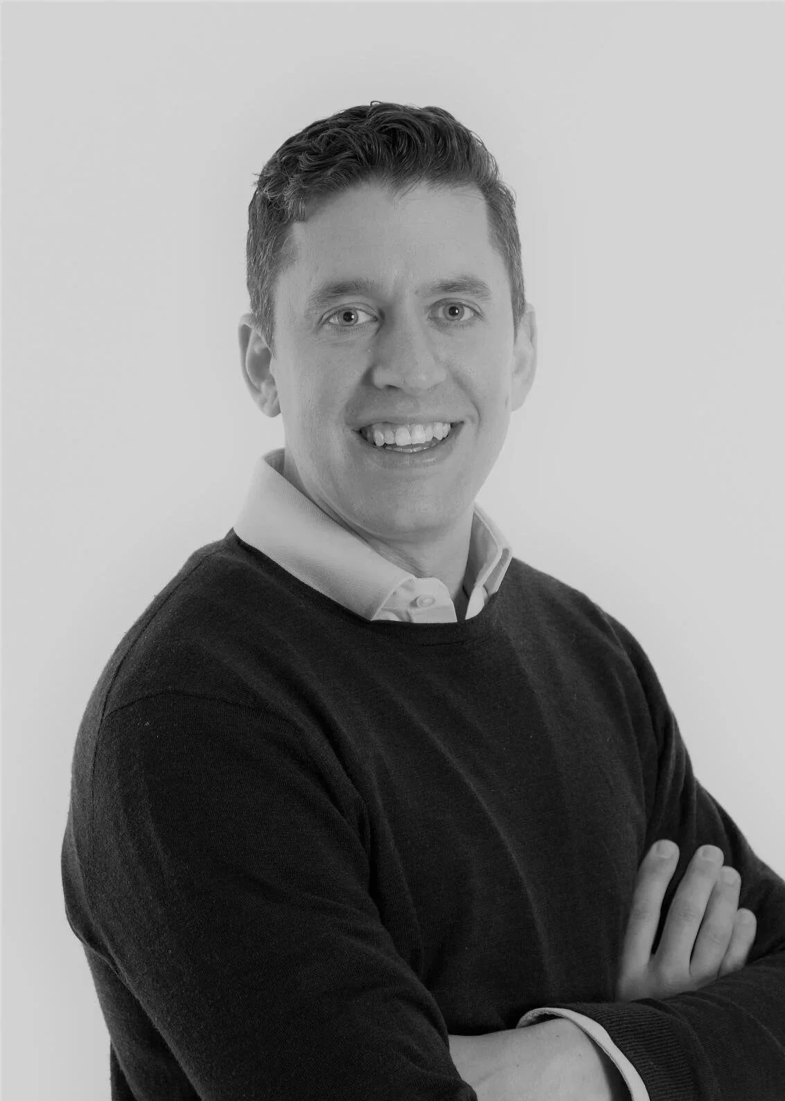 Black and white professional headshot of a man with short, wavy hair, wearing a collared shirt and sweater, smiling, with arms crossed, against a plain background.