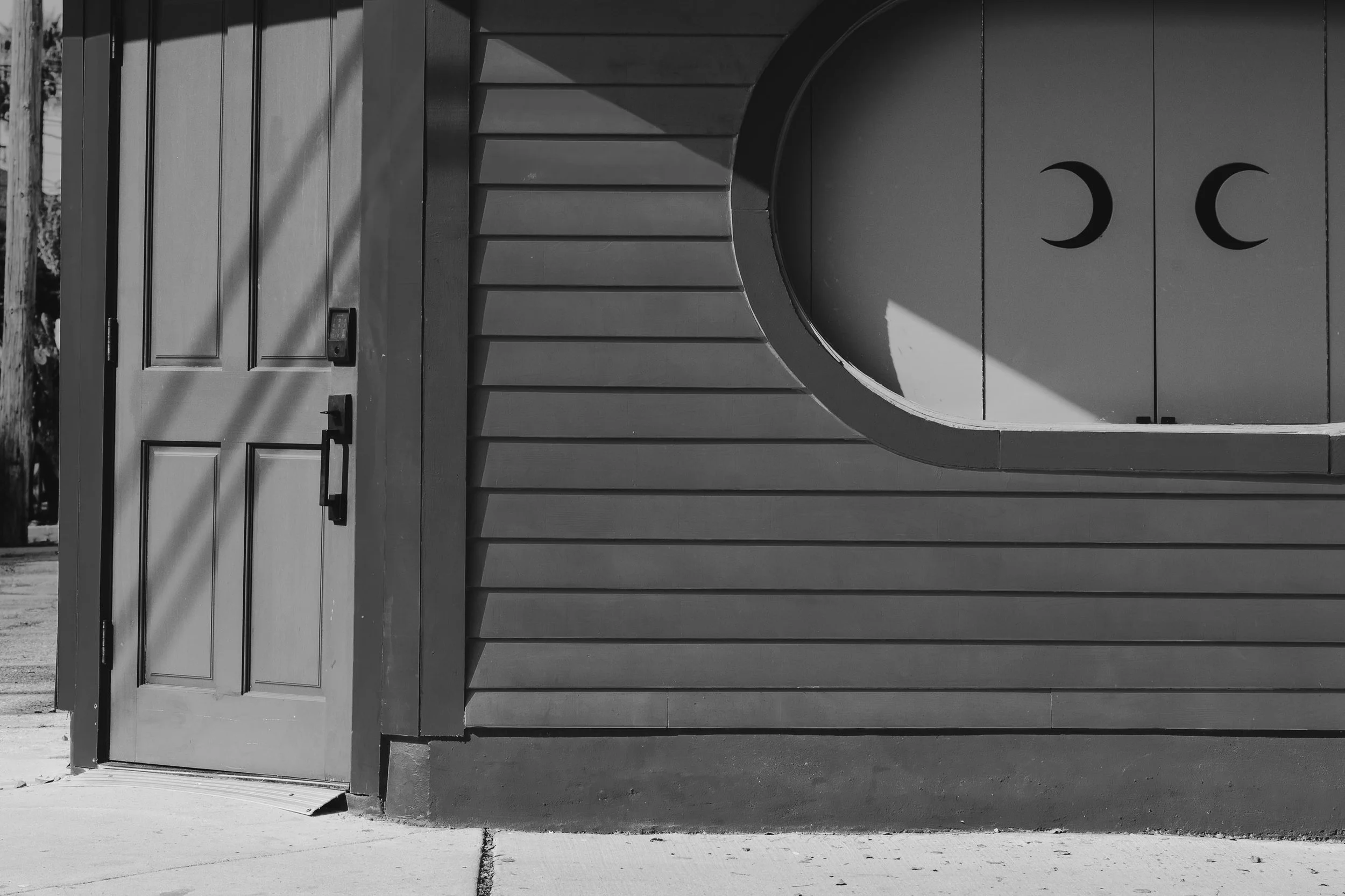 Waxing, Waning

Black-and-white photograph of a storefront with horizontal siding and a rounded window opening. The shutters feature mirrored crescent shapes, and a door stands slightly open at the left.