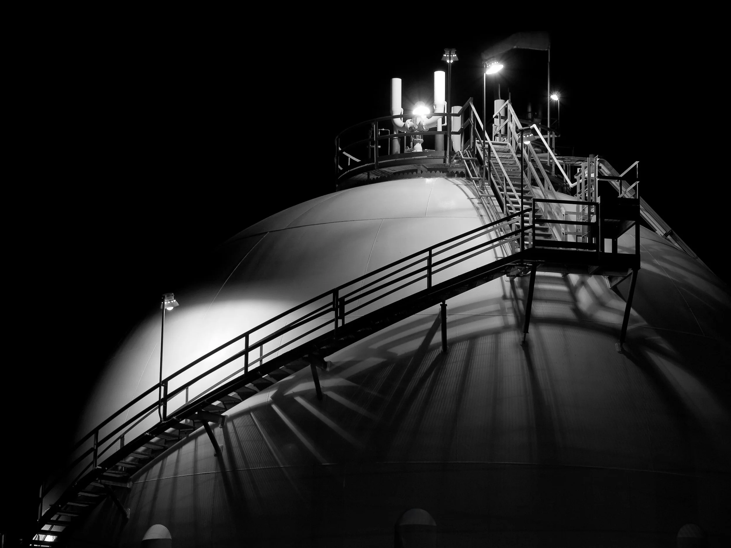 Non-Euclidean Projections

Black-and-white night photograph of a large spherical industrial storage tank with a curved metal staircase wrapping around its surface. Overhead lamps cast intersecting shadows across the smooth steel structure.