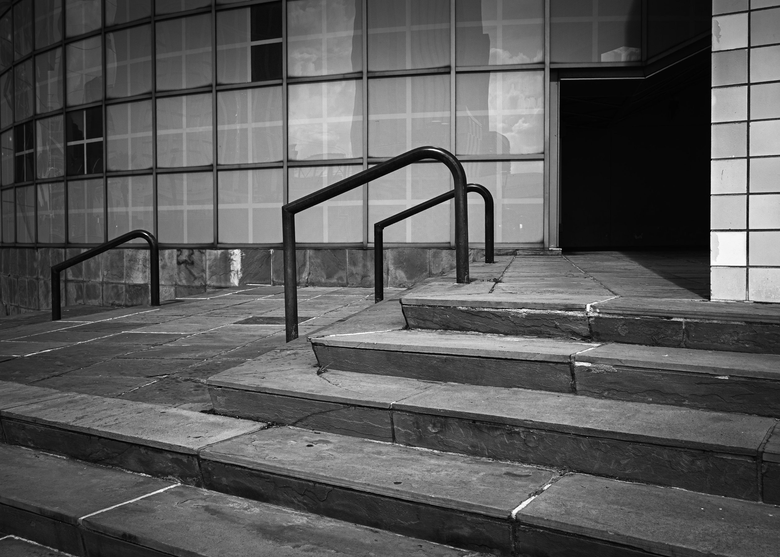 Black-and-white photograph of stone steps and metal handrails leading to the entrance of a modern building with tiled walls and a curved glass facade.