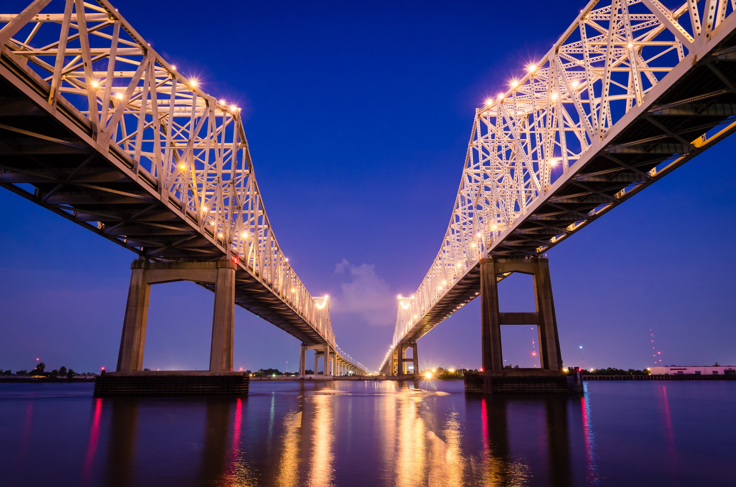 Unidentical Twins

Twilight photograph of the twin steel bridges of the Crescent City Connection spanning the Mississippi River. The illuminated trusses glow against a deep blue sky, with their lights reflected in the calm water below.