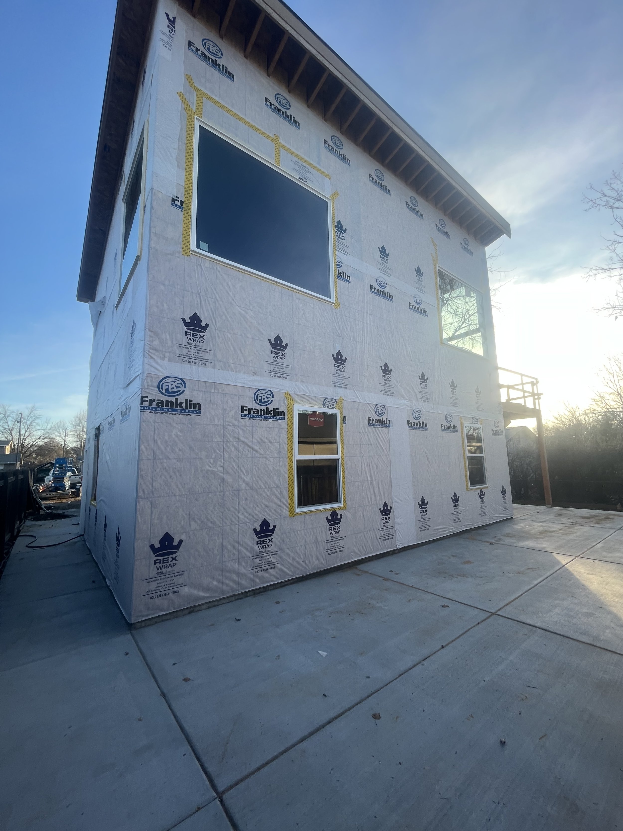 Exterior of a two-story house under construction with wrapped insulation, several windows, and a balcony on the second floor. The house is on a concrete slab foundation, and sunlight is illuminating the scene.